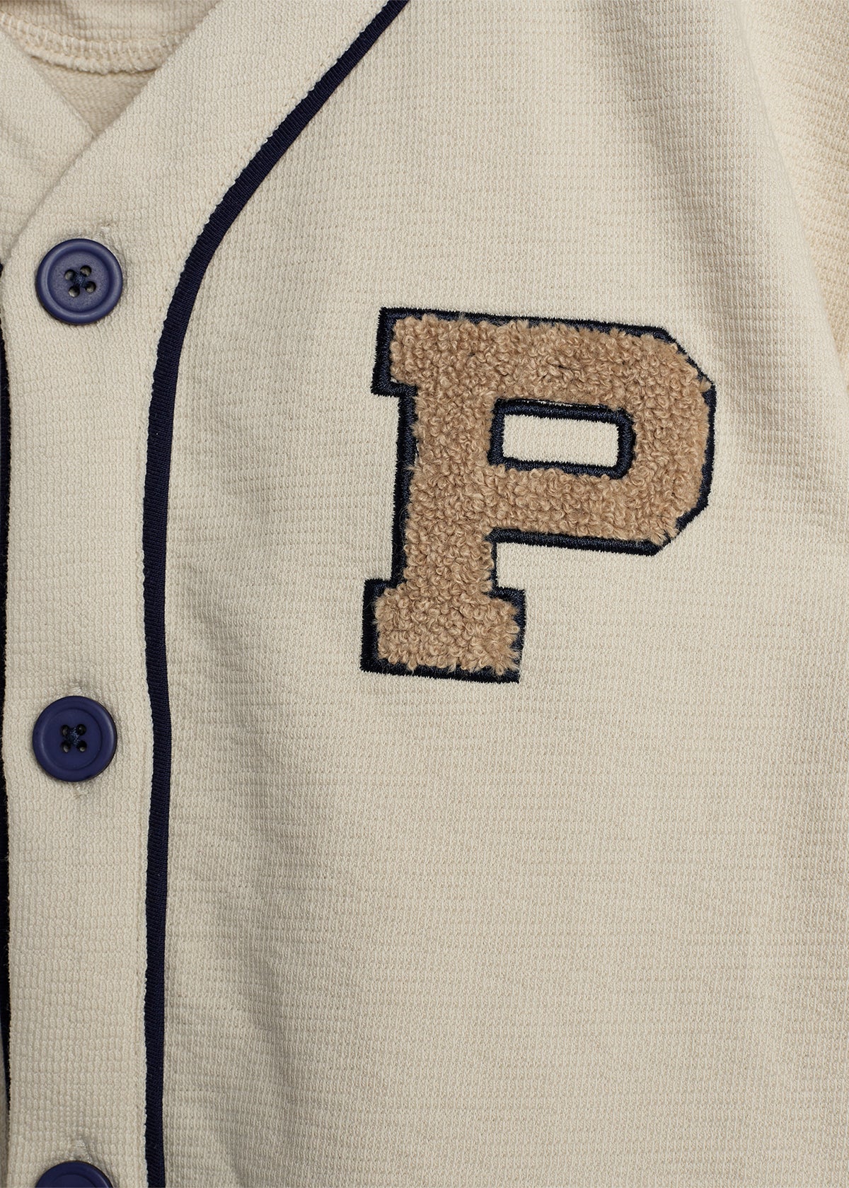 Eine beige Jacke mit dunkelblauen Knöpfen zeigt einen großen, applizierten Buchstaben "P" aus braunem Stoff. Die Jacke hat dunkelblaue Ränder und scheint sportlich gestaltet.