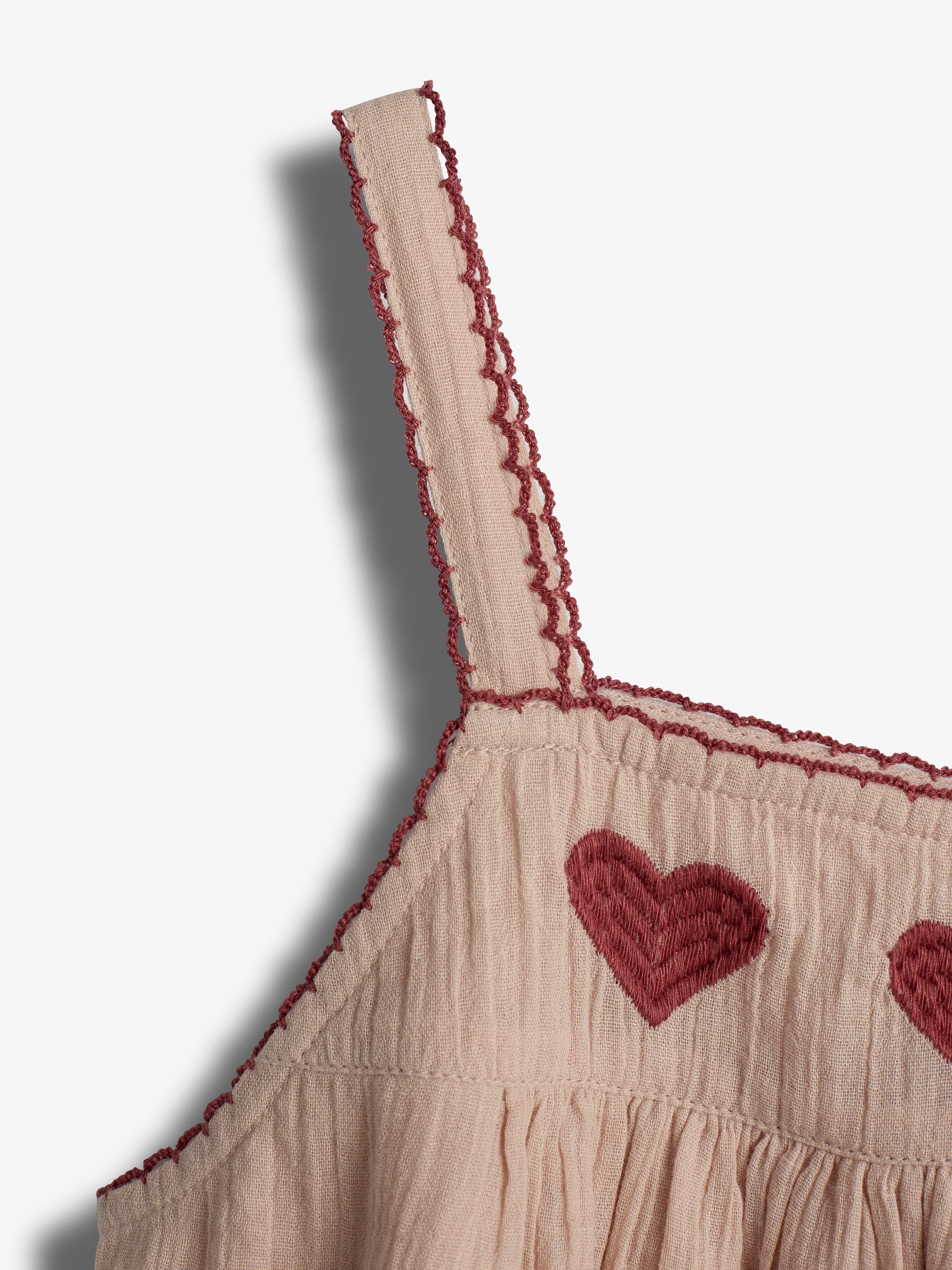 Ein beigefarbenes Kleid mit rot gestickten Herzmotiven auf der Vorderseite. Die Träger sind mit roten Ziernähten verziert, in einem minimalistischen hellgrauen Hintergrund.