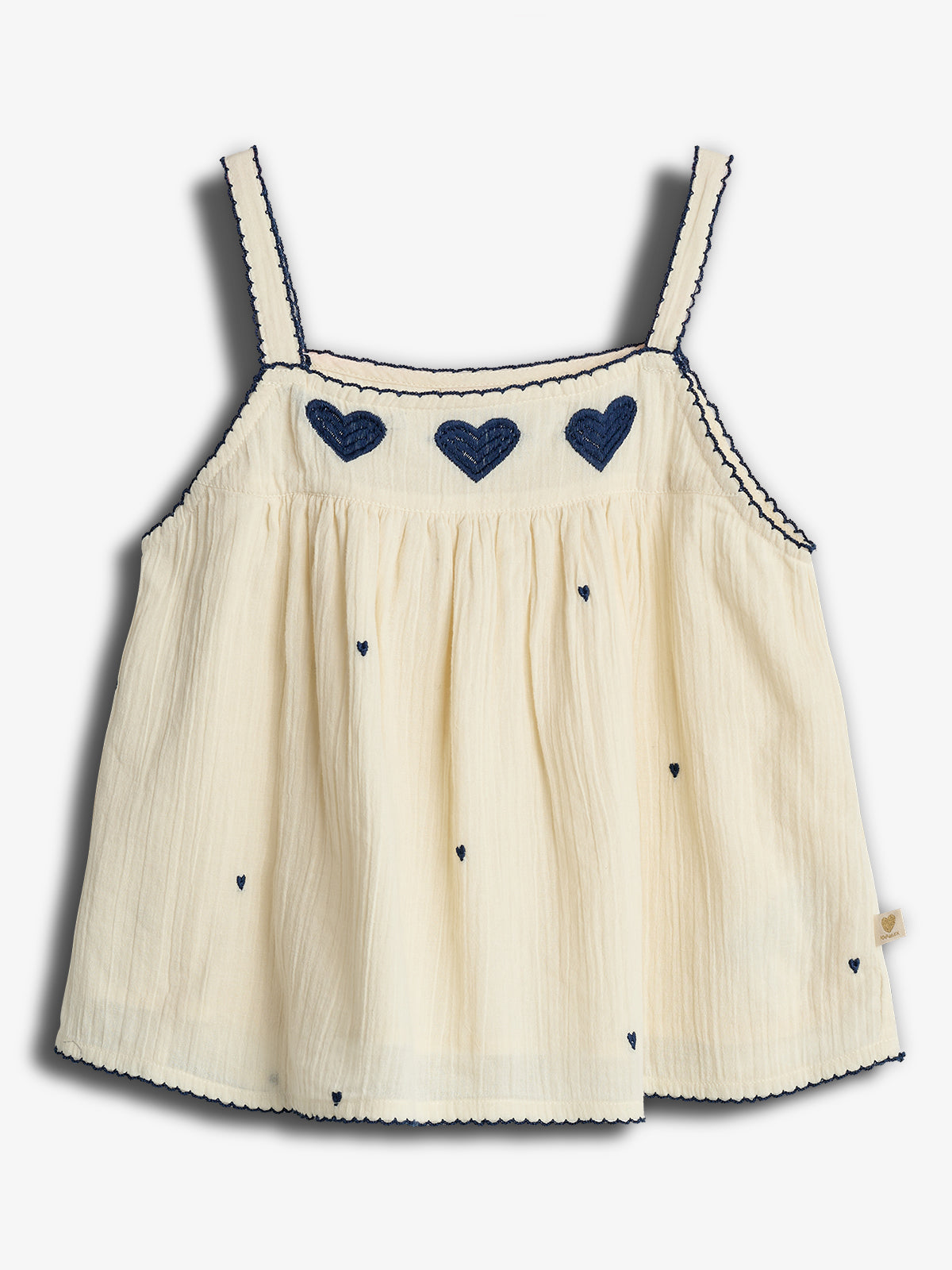 Ein cremefarbenes Kinderträgertop mit dunkelblauen herzförmigen Stickereien entlang des Brustbereichs. Die Träger sind mit blauen Details verziert. Es liegt auf einem weißen Hintergrund.