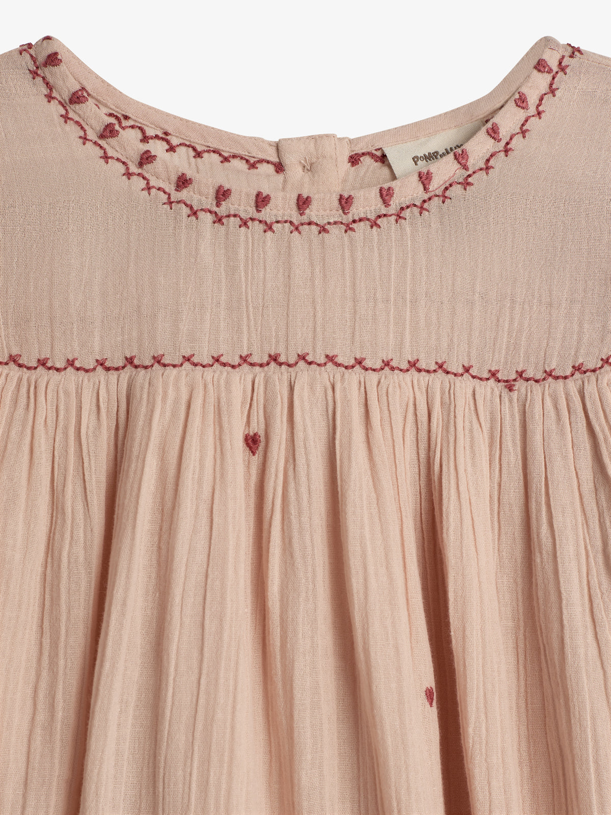 Ein hellrosa Kleid aus leichtem Stoff mit rötlichen Stickereien in Herzform am Kragen und entlang des Oberteils. Im Hintergrund ein Etikett mit dem Text: "Pompom".