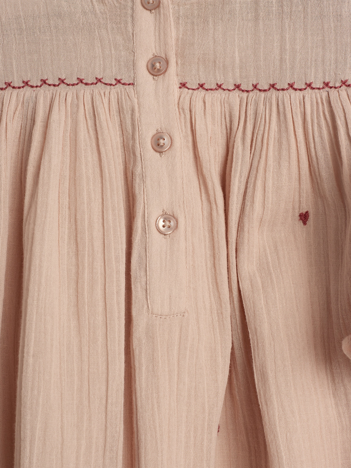 Beiges Kleid mit Knopfleiste, verziert mit rot gesticktem Muster entlang der Naht. Der Stoff ist leicht gerafft und zeigt eine dezente Struktur.