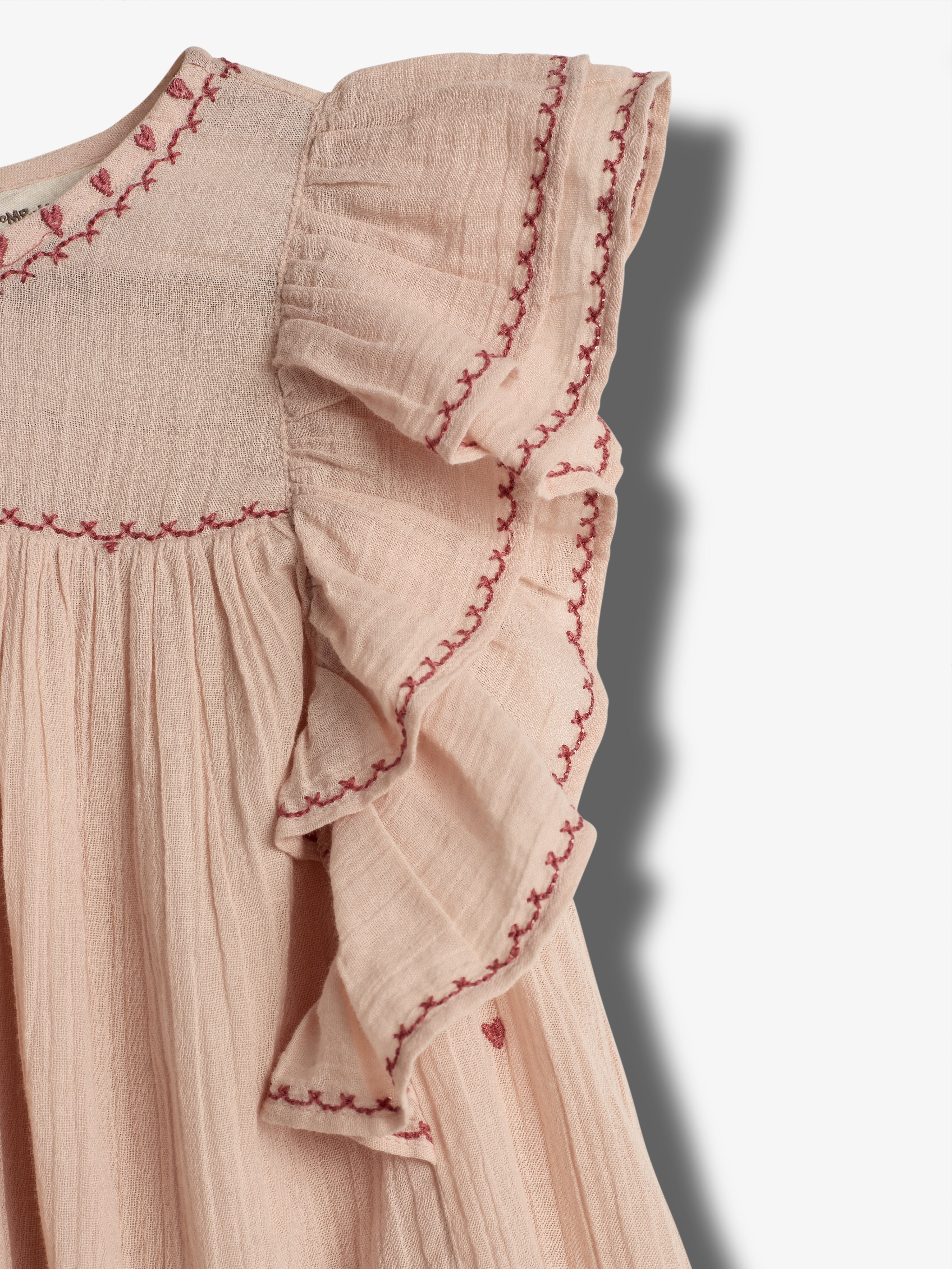 Ein rosa Kleid mit Rüschenärmel, verziert mit rot gestickten Mustern, hängt lose. Der Hintergrund ist schlicht und hell, wodurch das Kleid im Fokus steht.