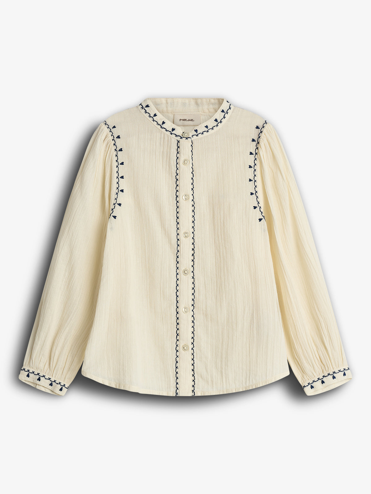 Eine cremefarbene Bluse mit blauer Stickerei entlang der Knopfleiste, am Kragen und den Ärmeln. Die Bluse hat lange Ärmel und ist auf einem weißen Hintergrund präsentiert.