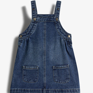 Jeanskleid mit Trägern und vier Knöpfen. Es hat zwei aufgesetzte Taschen vorne. Der Stoff ist dunkelblau, leicht verwaschen. Hintergrund ist ein neutraler, heller Ton.