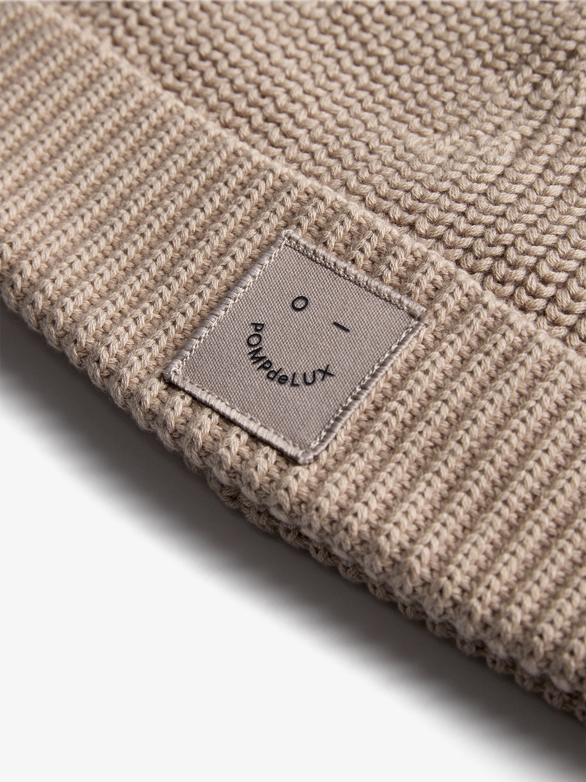Eine beige Strickmütze liegt flach. Ein Etikett zeigt ein stilisiertes Gesicht und den Text "POMPdeLUX". Die Oberfläche der Mütze ist grob gestrickt.