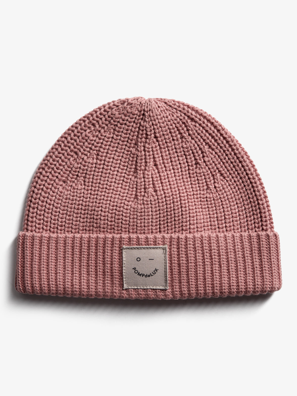 Eine rosa Strickmütze liegt auf einer weißen Fläche. Auf dem umgeschlagenen Rand befindet sich ein Etikett mit einem einfachen Gesicht und dem Text "POMPdeLUX".