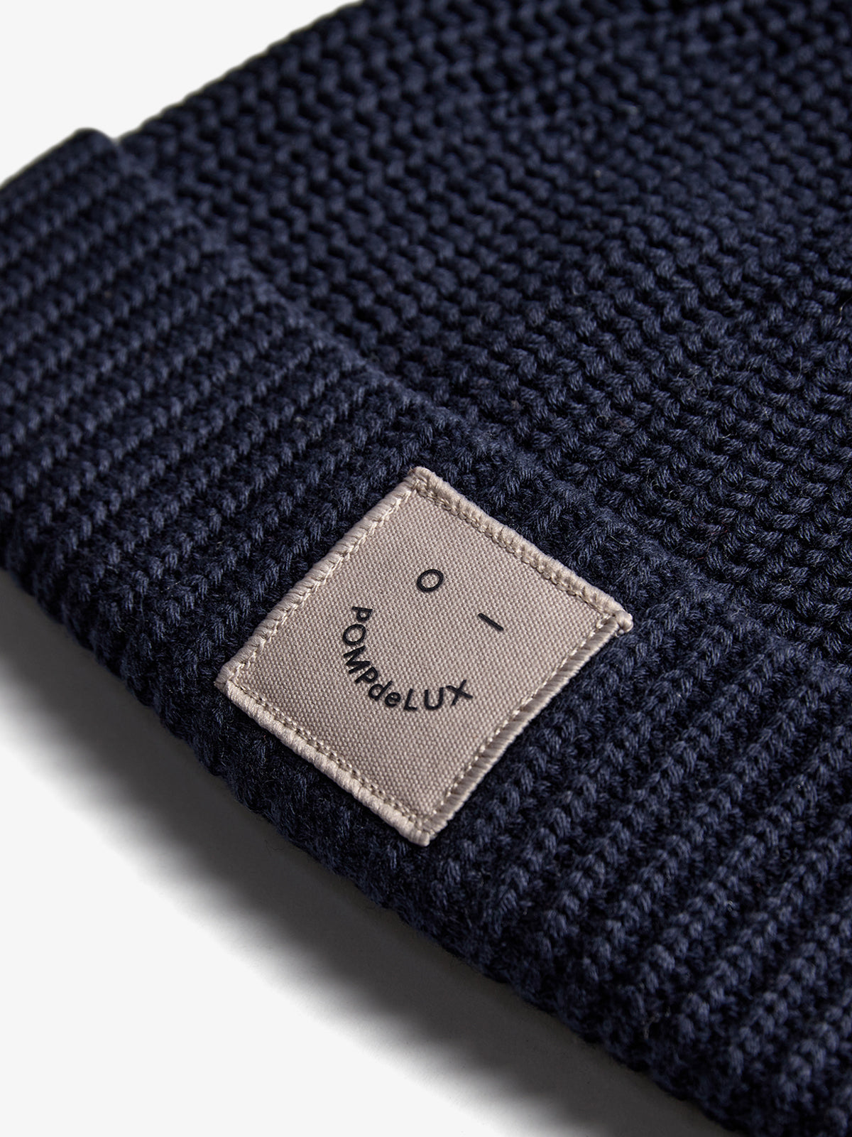 Eine dunkelblaue Strickmütze liegt unbewegt. Auf ihr ist ein beiges Etikett mit einem stilisierten Gesicht und dem Text "POMPdeLUX" aufgenäht. Der Hintergrund ist weiß.