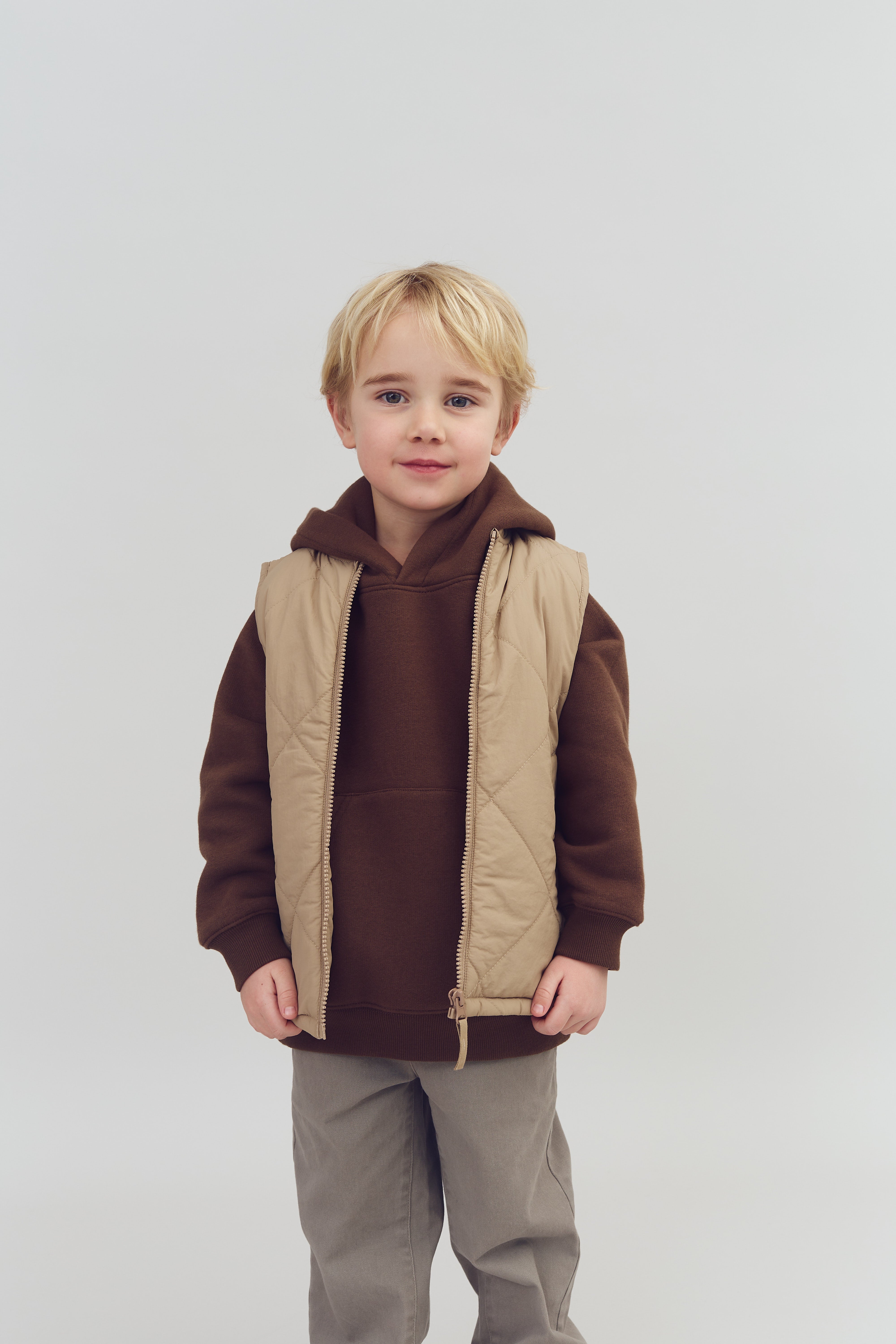 Ein Kind trägt eine beige Weste über einem braunen Kapuzenpullover und graue Hosen. Es steht vor einem neutralen, hellen Hintergrund.