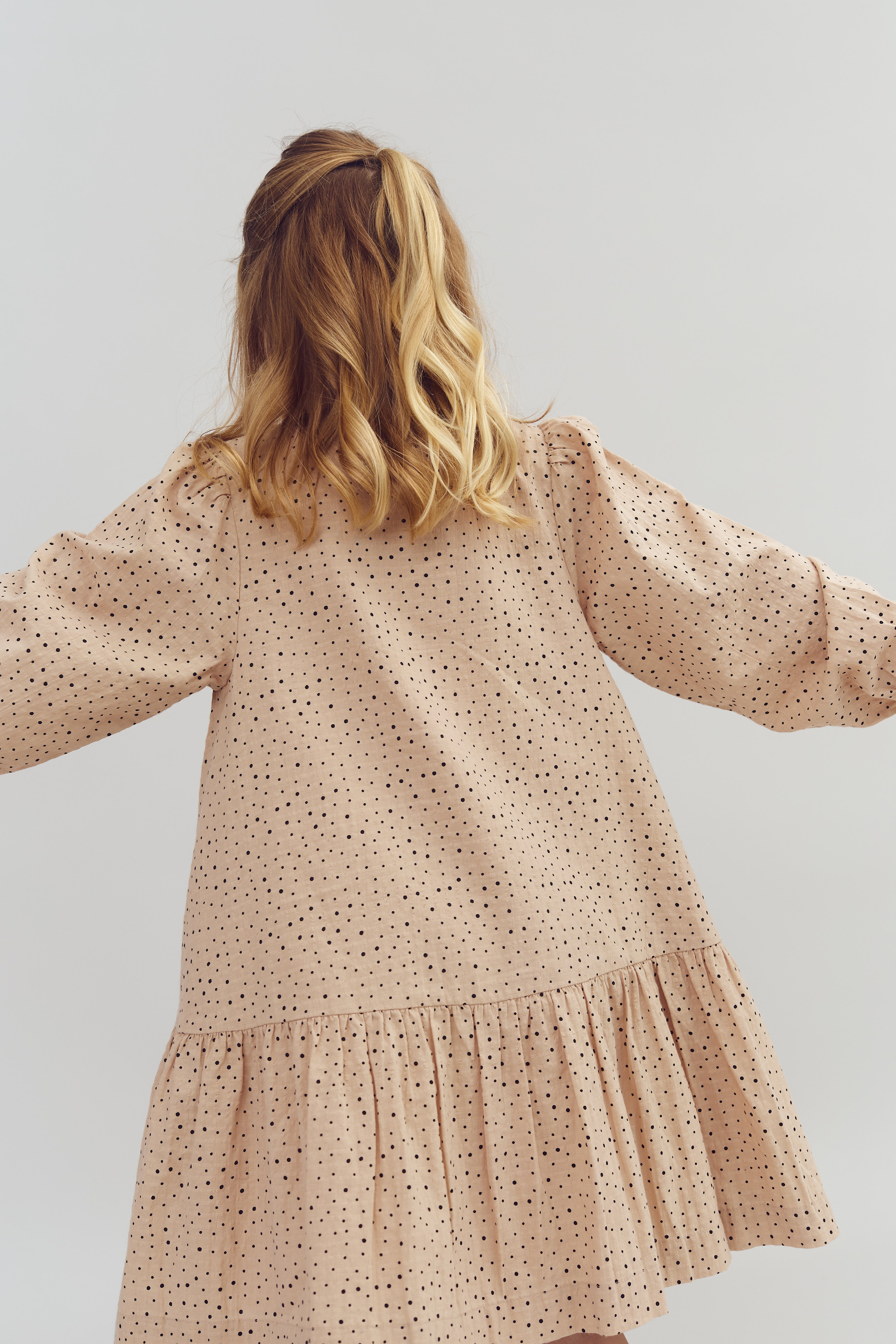 Eine Person von hinten, trägt ein beigefarbenes Kleid mit schwarzen Punkten. Die Haare sind blond und gewellt, der Hintergrund ist neutral grau.