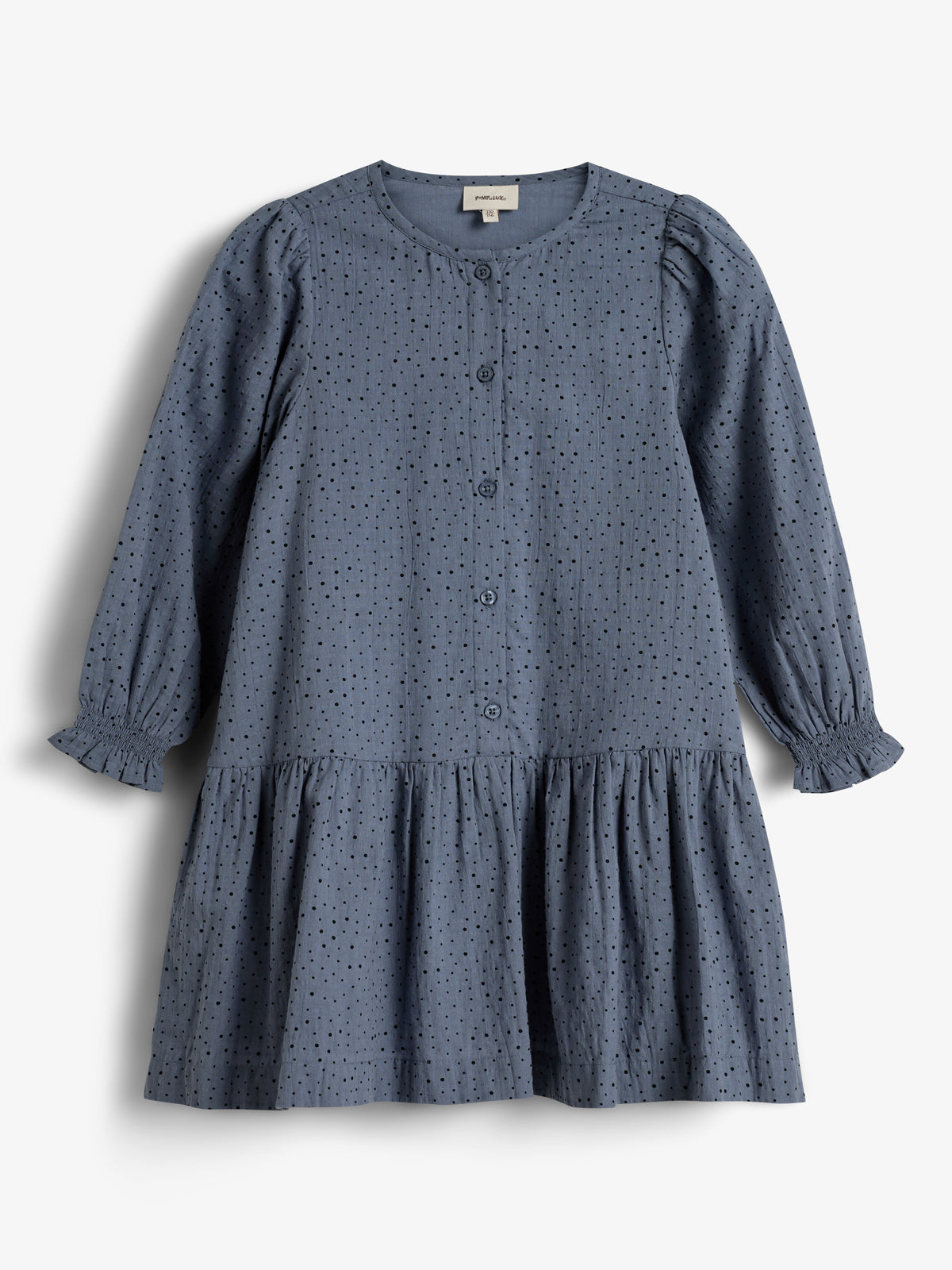 Ein blaues, gepunktetes Kleid hängt lose, mit langen Ärmeln und Knopfleiste auf weißem Hintergrund. Der Saum ist gerafft, und der Stoff wirkt leicht strukturiert.