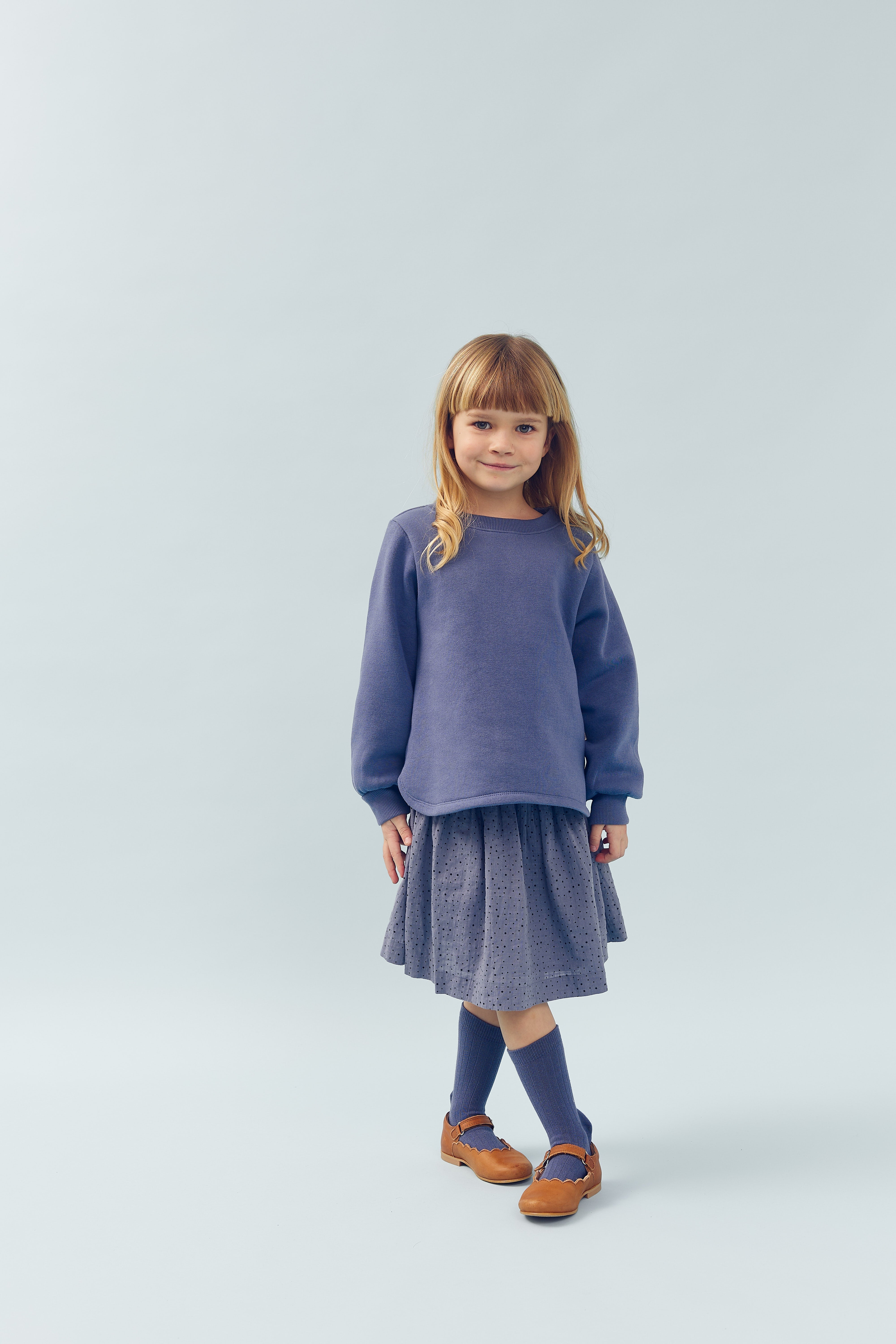 Ein Kind, das einen blauen Pullover und einen gepunkteten Rock trägt, steht lächelnd in einem hellen, neutralen Raum. Sie trägt blaue Kniestrümpfe und braune Mary-Jane-Schuhe.
