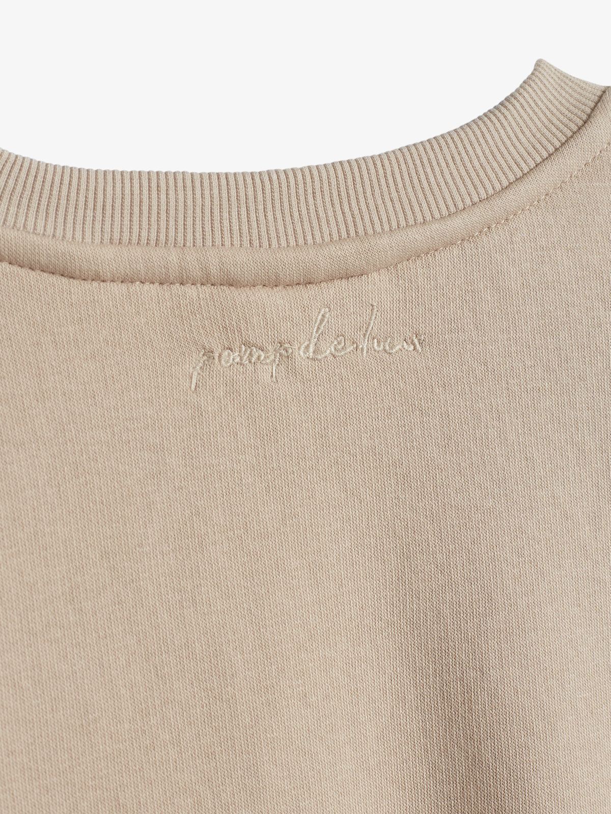 Ein beigefarbenes Sweatshirt zeigt oben nahe dem Kragen die gestickte Schrift "poupele huis", umgeben von geripptem Stoff in einem neutralen, minimalistischen Stil.