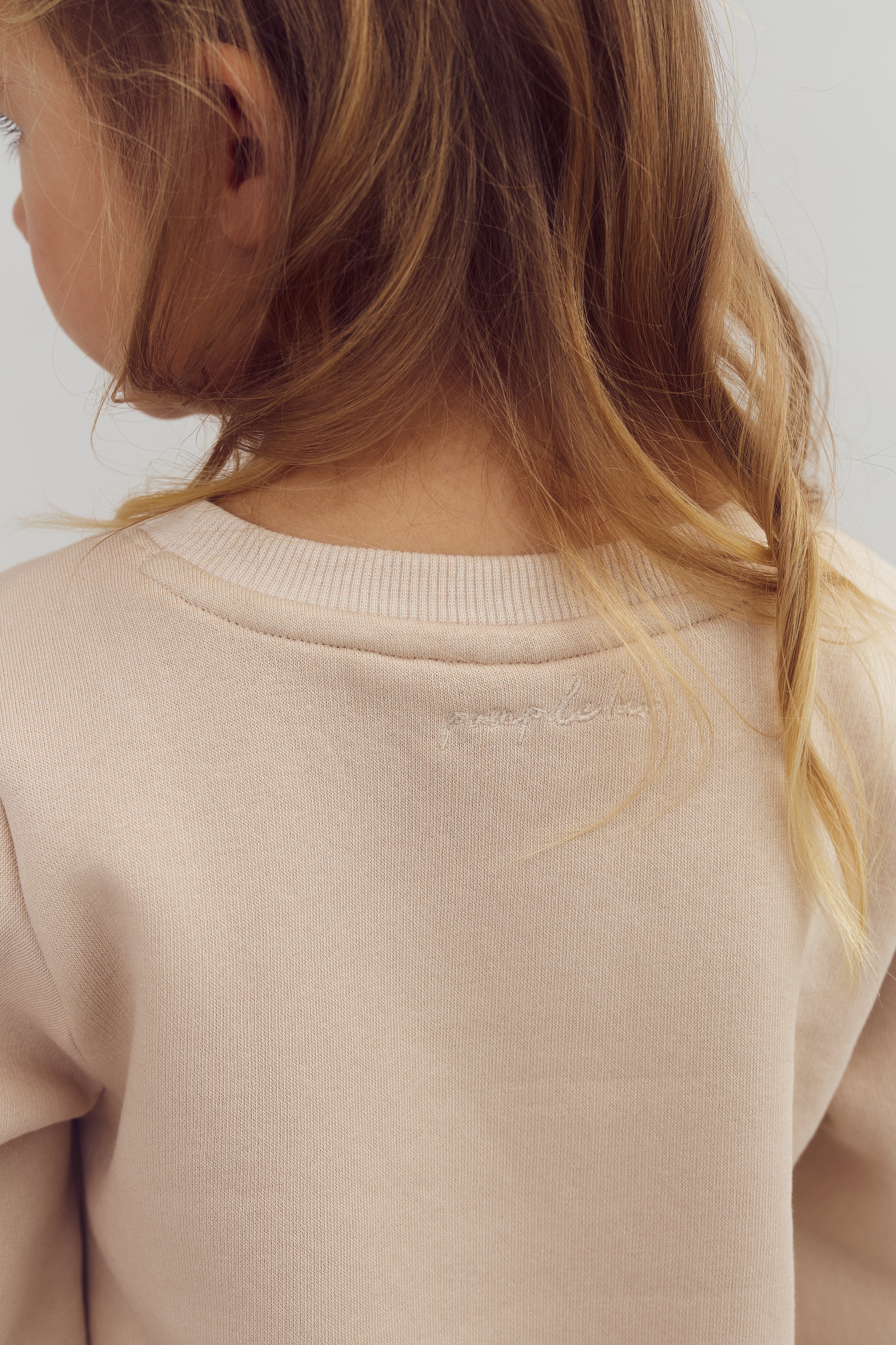 Ein Kind präsentiert den Rücken in einem beigefarbenen Pullover, das Gesicht zur Seite gedreht, in einem schlichten, hellen Raum. Die Aufschrift auf dem Pullover: "peaceful soul".