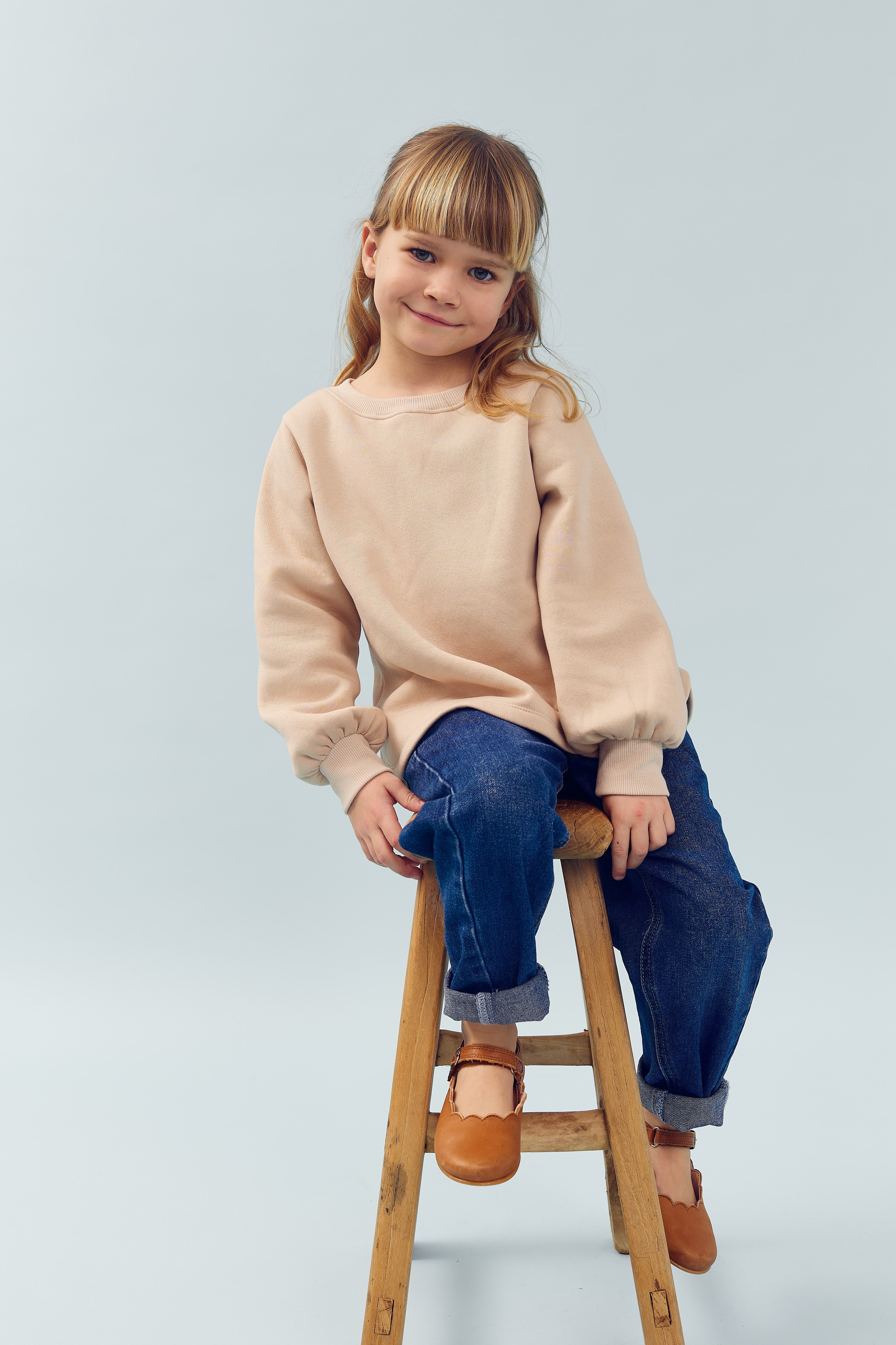 Kind auf einem Holzstuhl, lächelt leicht, trägt beigen Pullover und blaue Jeans, sitzt in einem neutralen, hellen Raum. Keine Textelemente vorhanden.