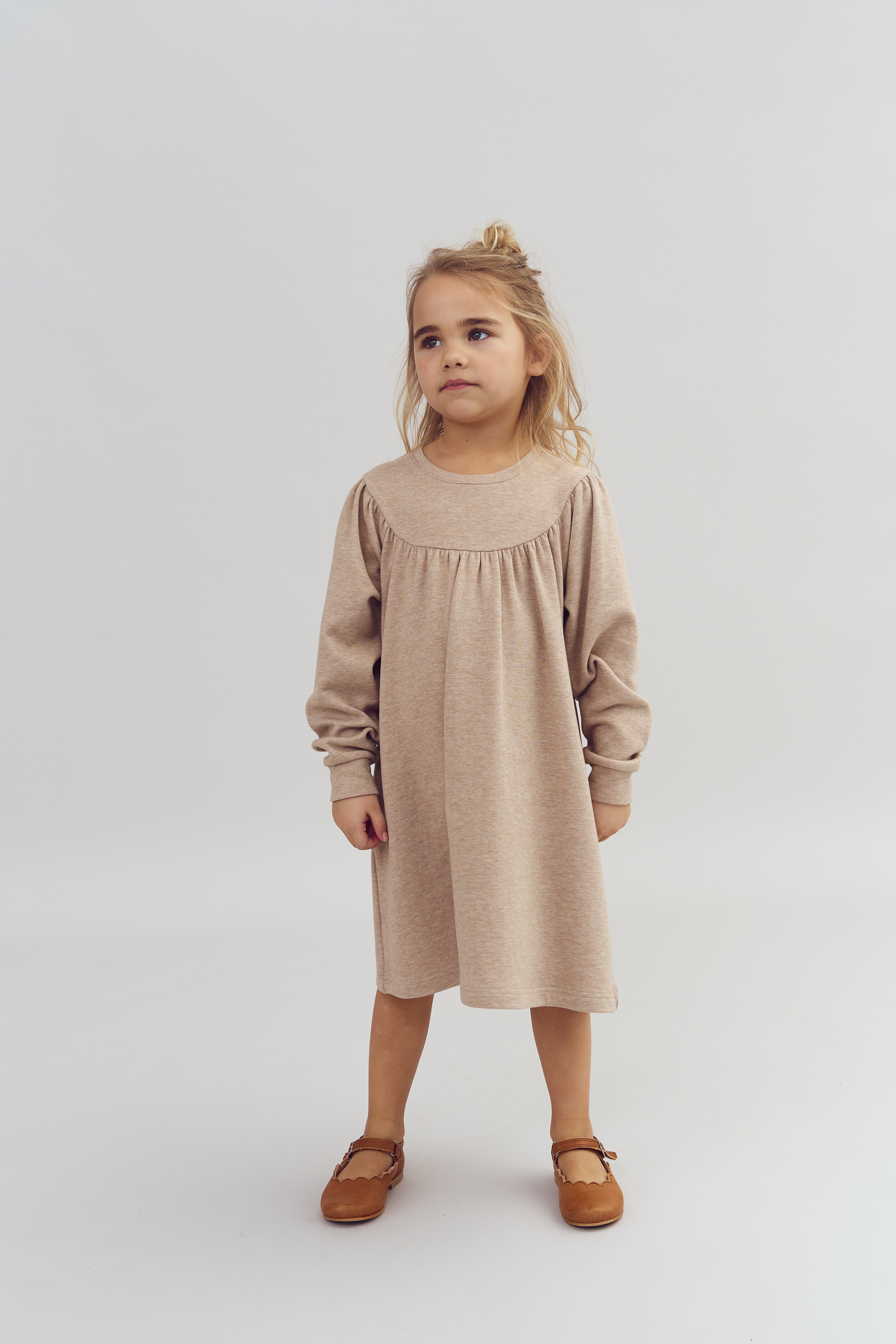 Ein Kind steht ruhig in einem beigen Kleid und braunen Schuhen, in einem hellen, neutralen Raum, während es nachdenklich in die Ferne blickt.