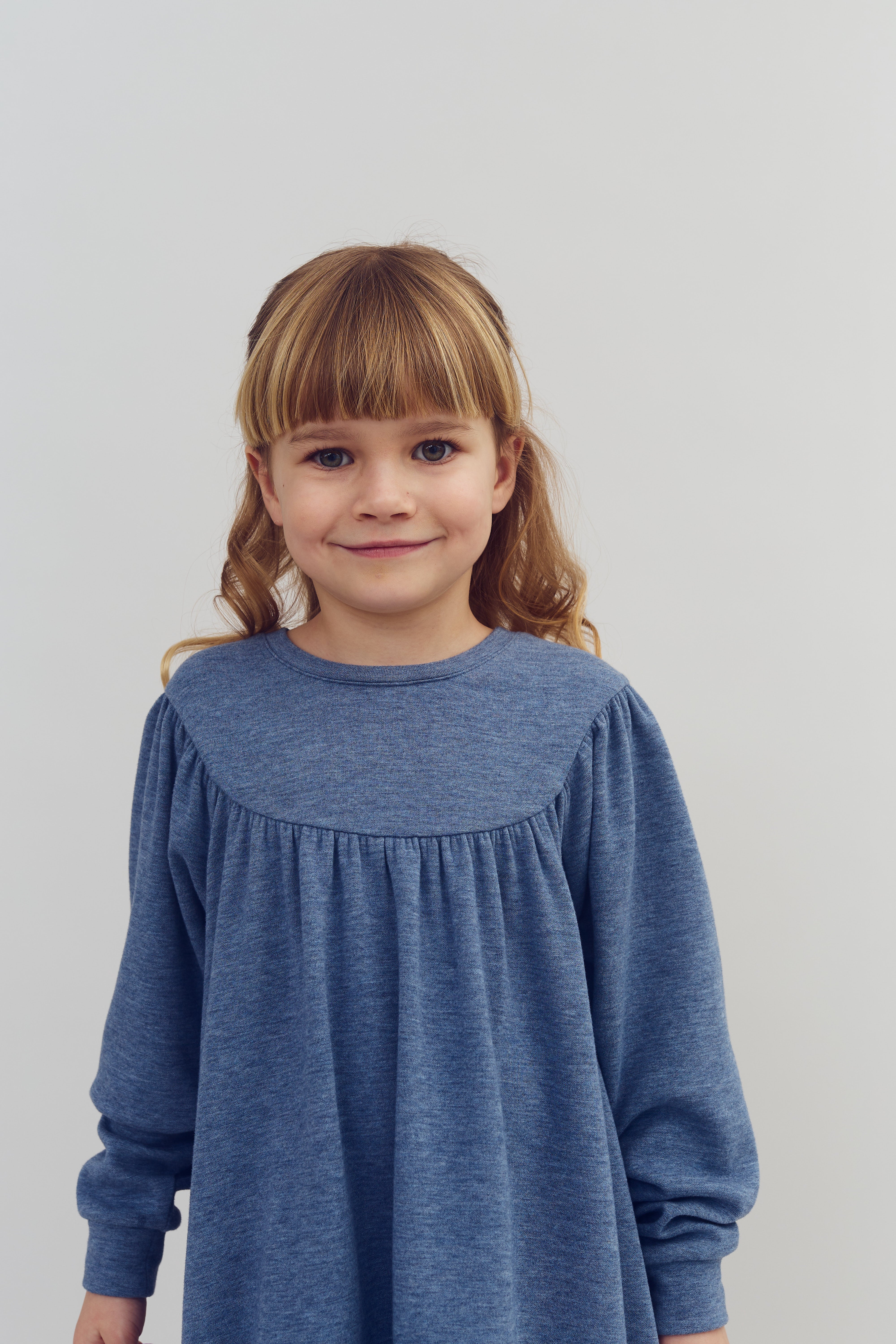 Ein Kind trägt ein blaues Kleid, lächelt in die Kamera und steht vor einem neutralen, hellen Hintergrund.