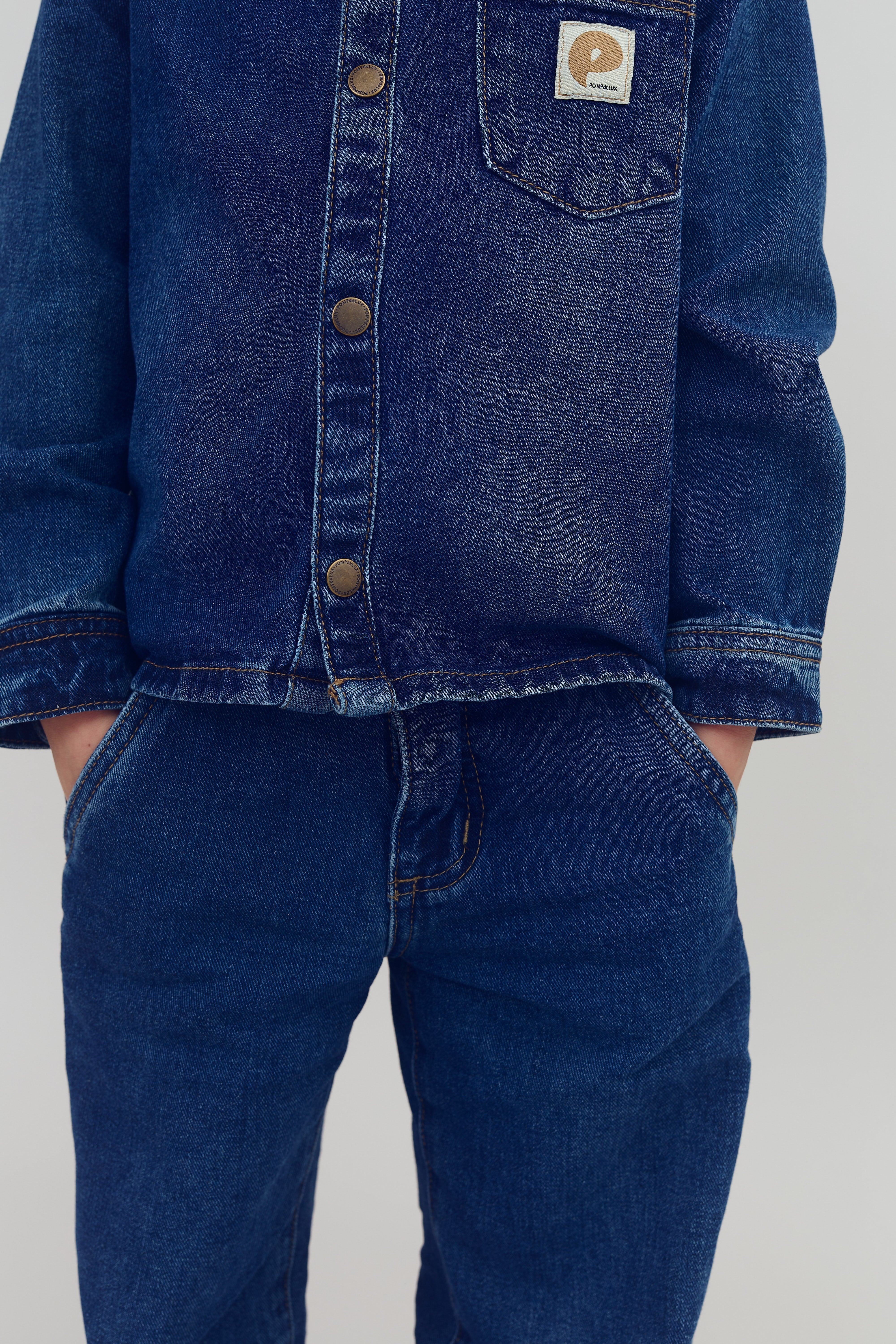 Jeansjacke mit Knöpfen und Brusttasche, die ein aufgenähtes Logo zeigt. Hände stecken in den Hosentaschen der passenden Jeanshose. Der Hintergrund ist schlicht.
 
 Text: "POLARN O. PYRET"