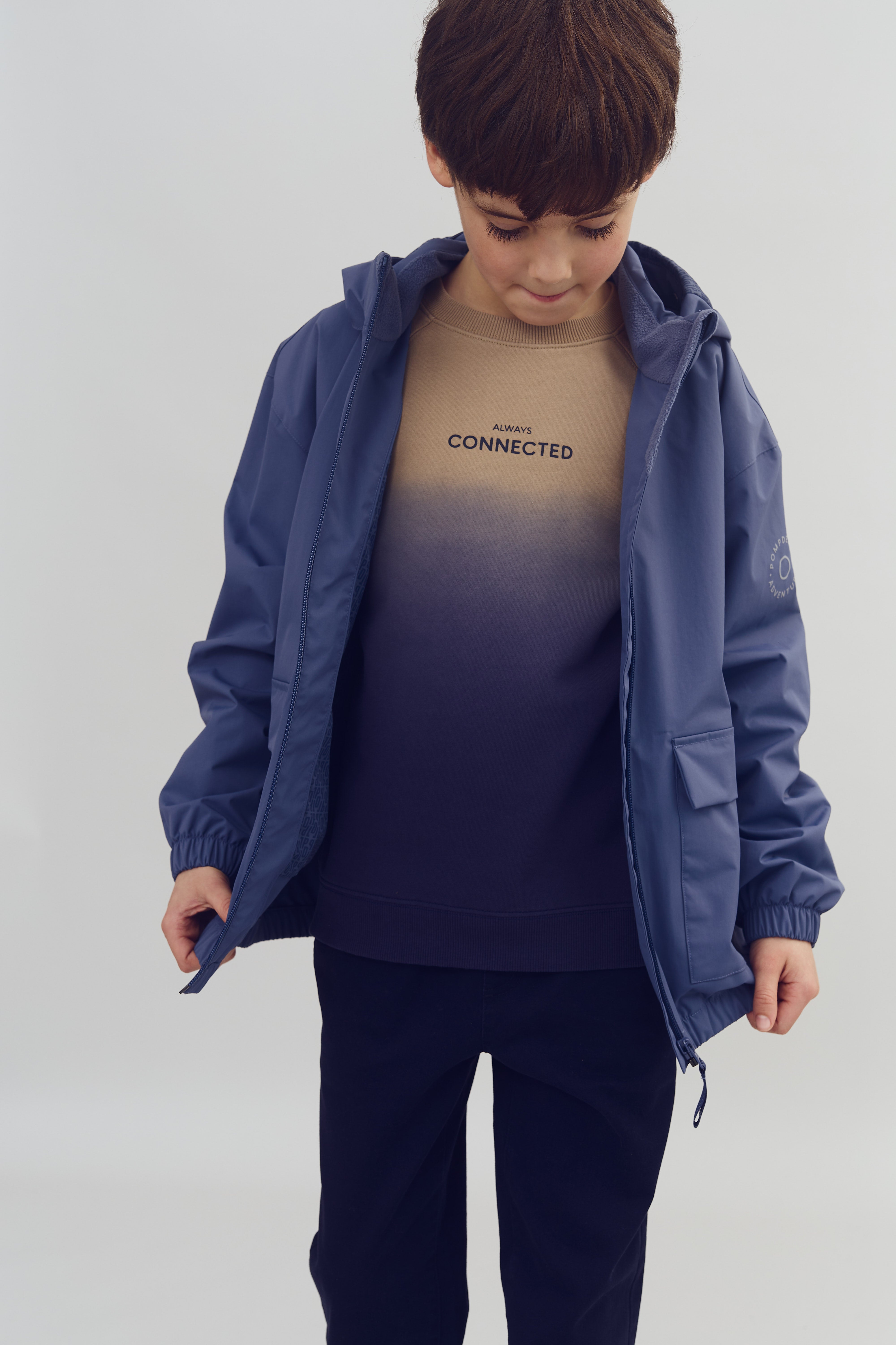 Ein Junge trägt eine blaue Jacke und ein Oberteil mit der Aufschrift "ALWAYS CONNECTED". Er schaut nach unten. Der Hintergrund ist neutral und schlicht.