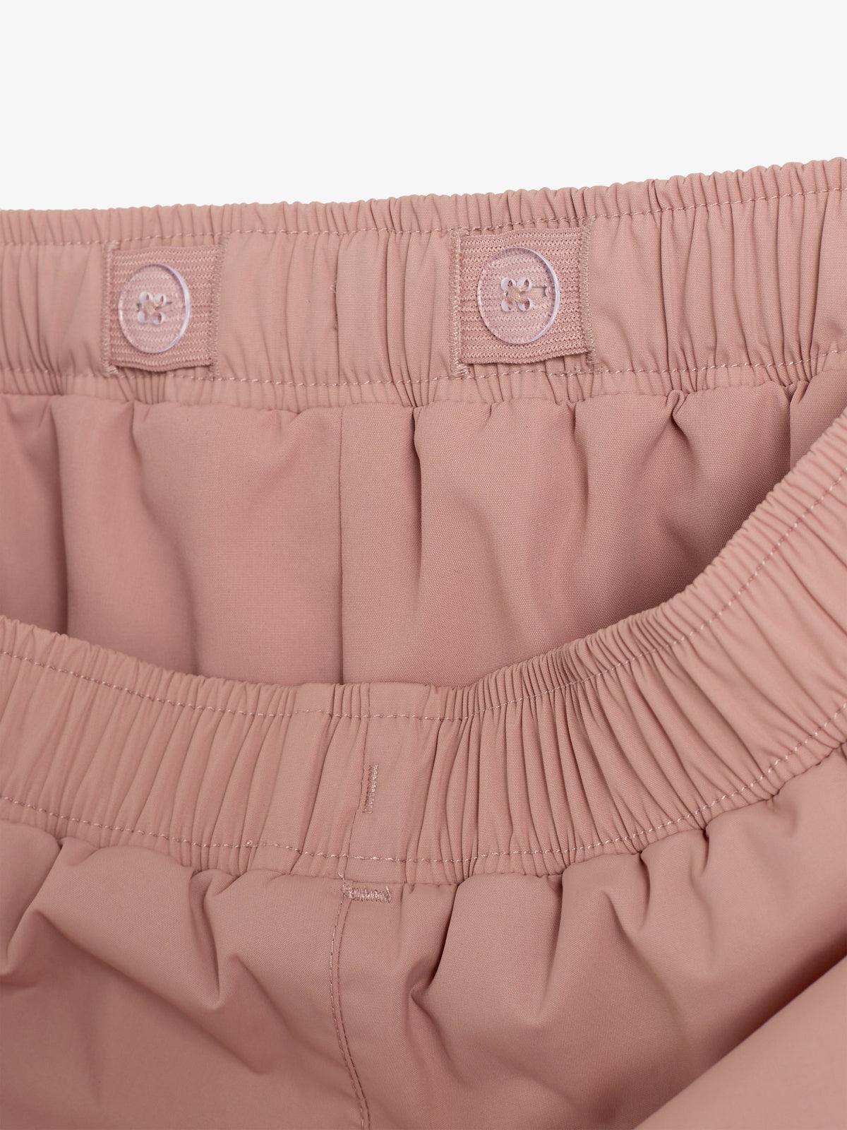 Der Bund einer rosa Hose mit elastischem Gummizug und Knöpfen auf der Vorderseite zeigt eine sorgfältige Verarbeitung. Background ist ein neutraler, einfarbiger Hintergrund.