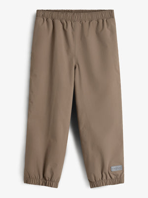 Eine braune, knöchellange Hose mit elastischem Bund und Beinabschlüssen liegt flach ausgebreitet auf einem weißen Hintergrund. Ein graues Etikett mit der Aufschrift "POM POMPS" befindet sich am unteren Rand.