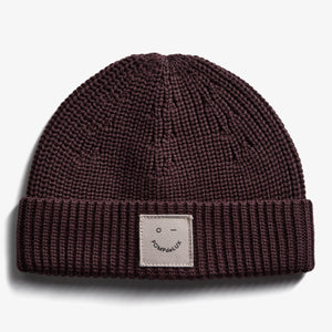 Eine gestrickte, dunkelbraune Mütze mit einem umgeschlagenen Rand. Auf dem Umschlag gibt es ein beiges Label mit einem minimalistischen Gesicht und dem Text "POMPdeLUX". Hintergrund in schlichtem Weiß.