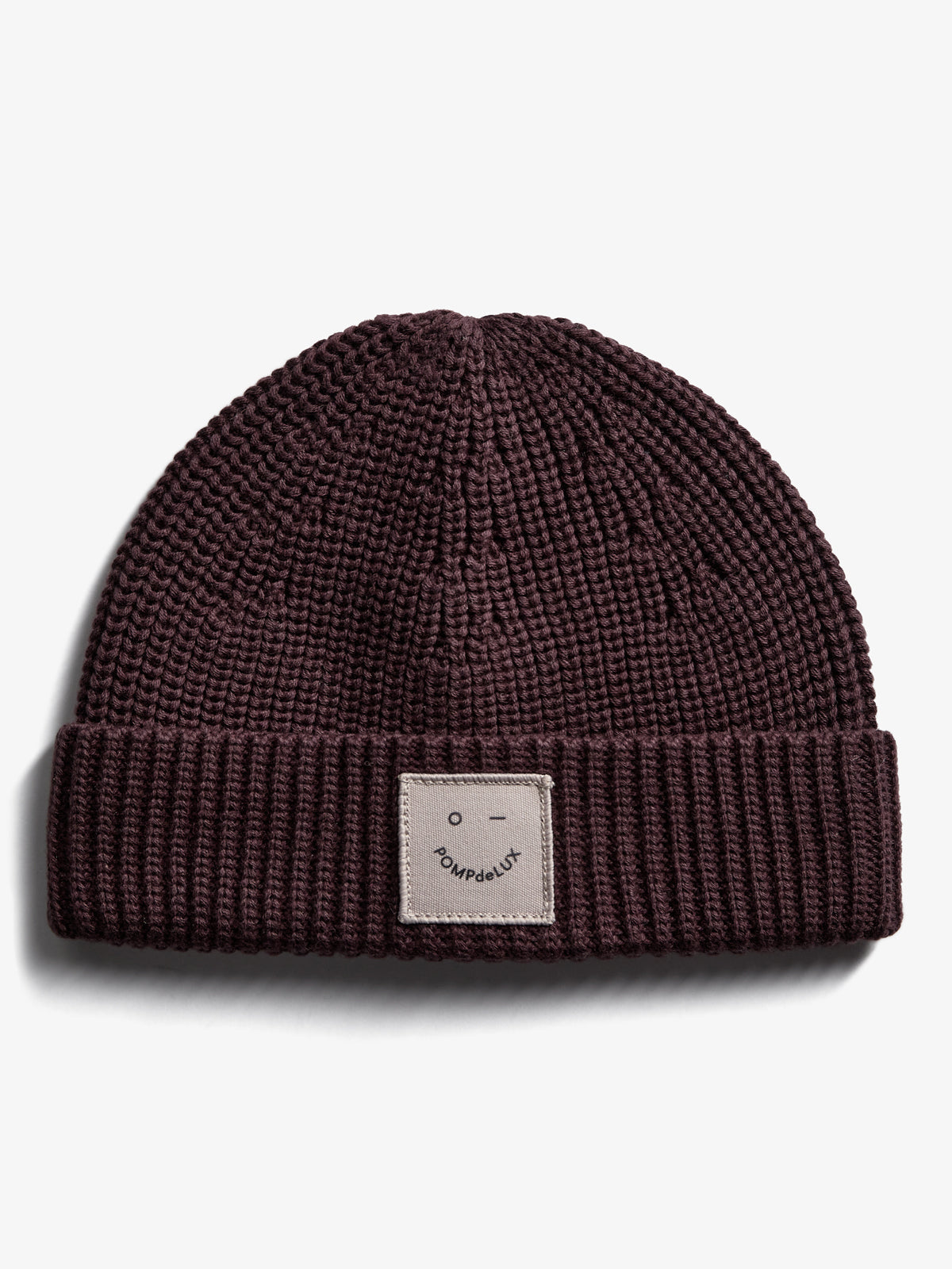 Eine gestrickte, dunkelbraune Mütze mit einem umgeschlagenen Rand. Auf dem Umschlag gibt es ein beiges Label mit einem minimalistischen Gesicht und dem Text "POMPdeLUX". Hintergrund in schlichtem Weiß.