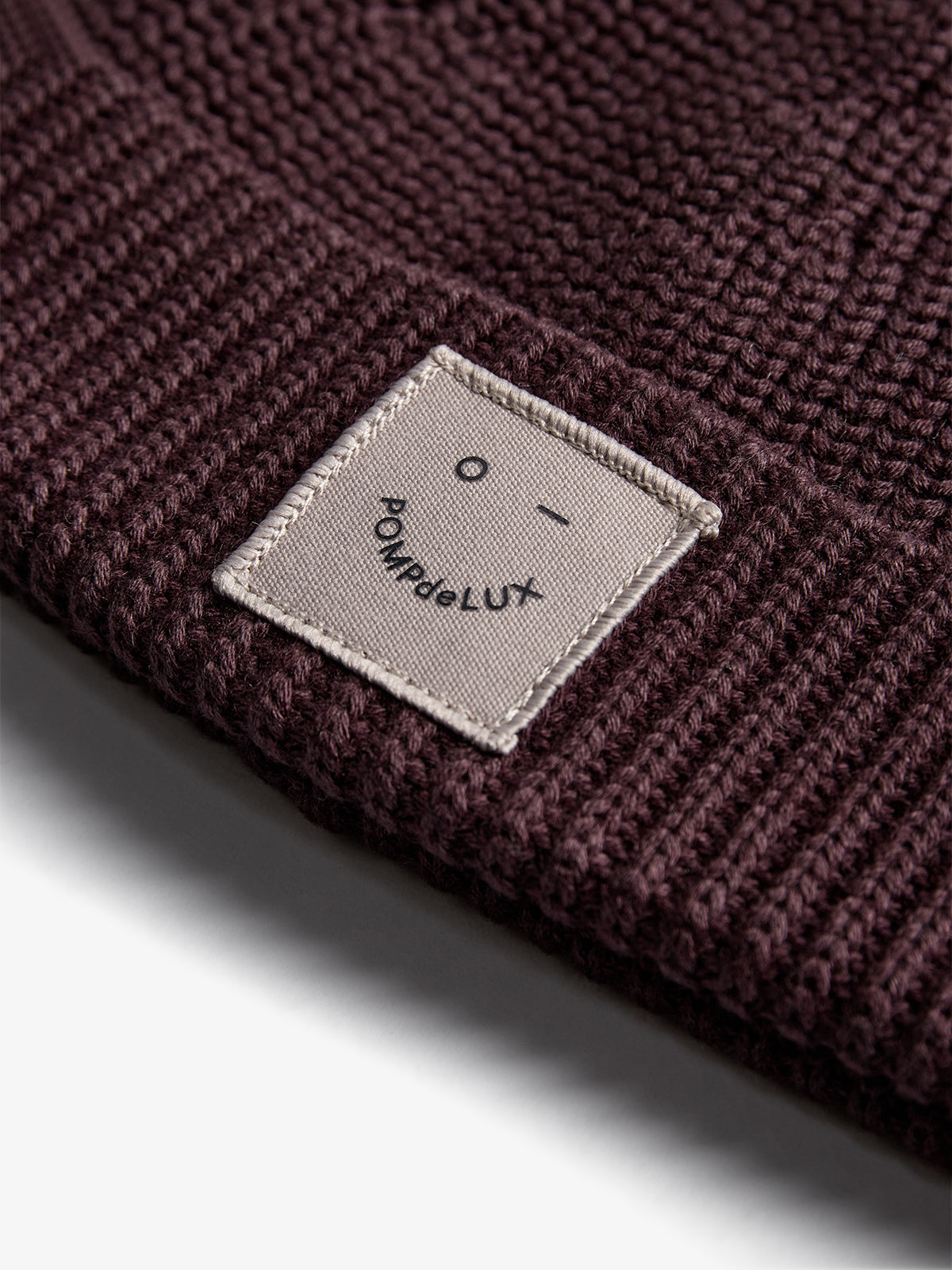 Braune Strickmütze zeigt ein eingenähtes Etikett mit einem lächelnden Gesicht und dem Text "POMPdeLUX". Liegt in einer beleuchteten, einfachen Umgebung.