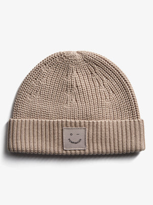 Eine beige Strickmütze mit umgeschlagener Krempe, auf der ein kleines Etikett mit einem stilisierten Gesicht und der Aufschrift "POMPdeLUX" angebracht ist, liegt auf weißem Hintergrund.
