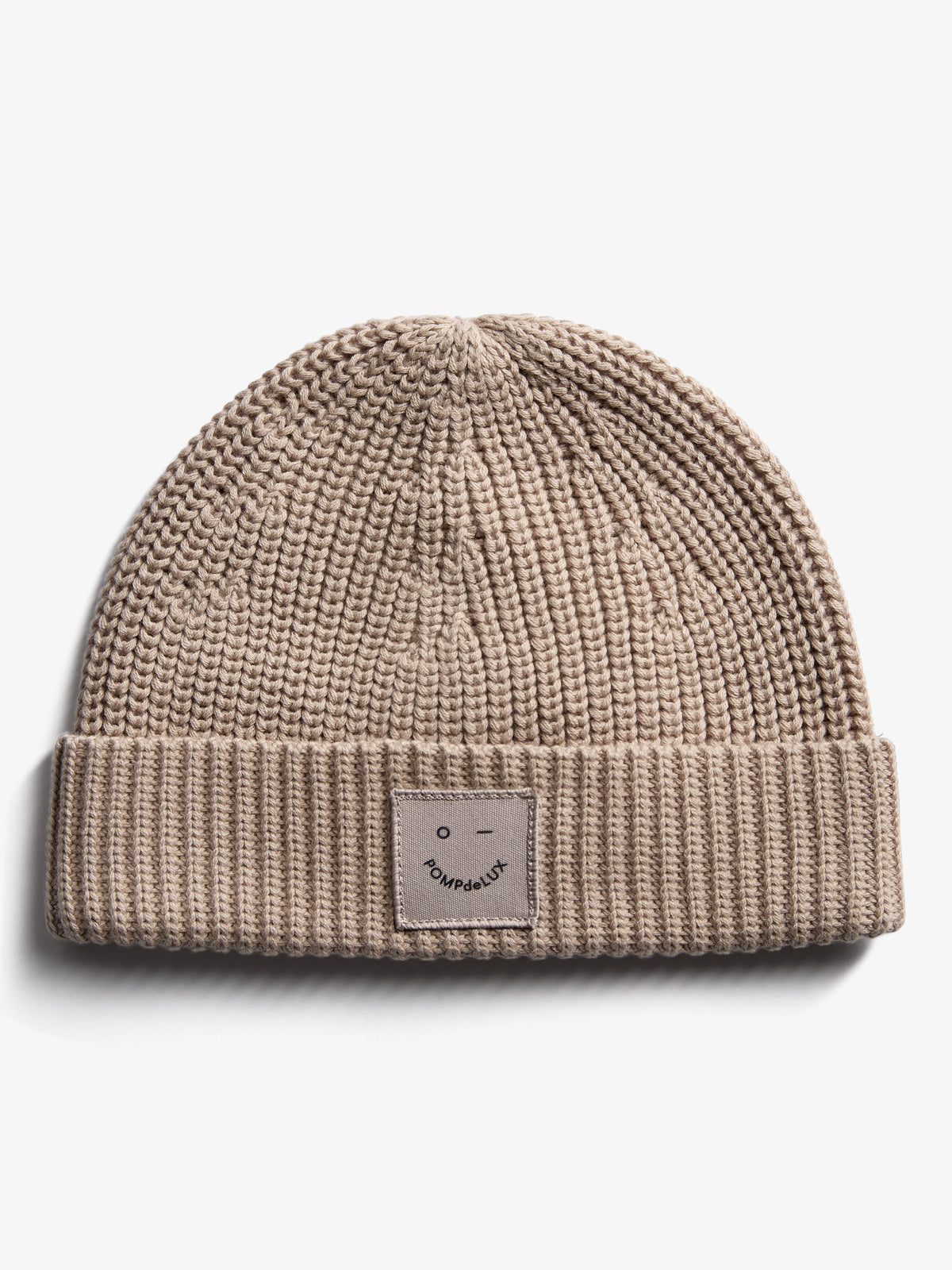 Eine beige Strickmütze mit umgeschlagener Krempe, auf der ein kleines Etikett mit einem stilisierten Gesicht und der Aufschrift "POMPdeLUX" angebracht ist, liegt auf weißem Hintergrund.
