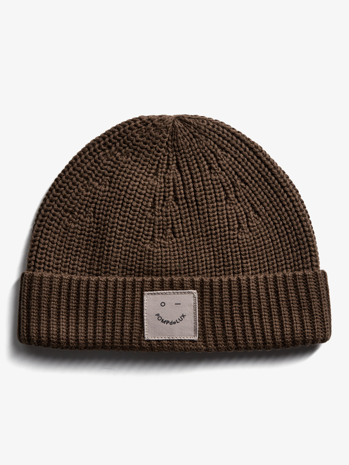 Eine braune Strickmütze liegt flach auf einem weißen Hintergrund. Auf der umgeschlagenen Krempe befindet sich ein quadratisches Etikett mit einem Smiley und dem Text "POMPdeLUX".