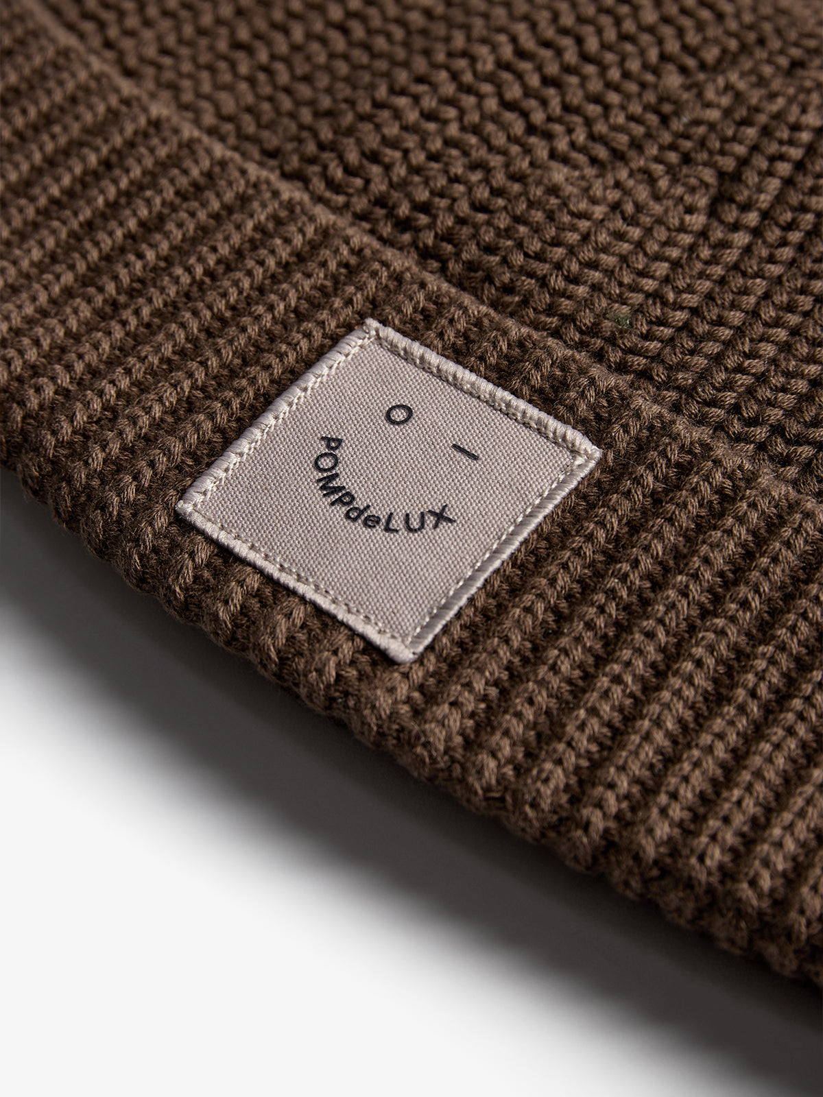 Eine braune Strickmütze mit einem aufgenähten quadratischen Etikett, das einen Smiley und den Text "POMPdeLUX" zeigt; die Mütze liegt auf einer neutralen Oberfläche.