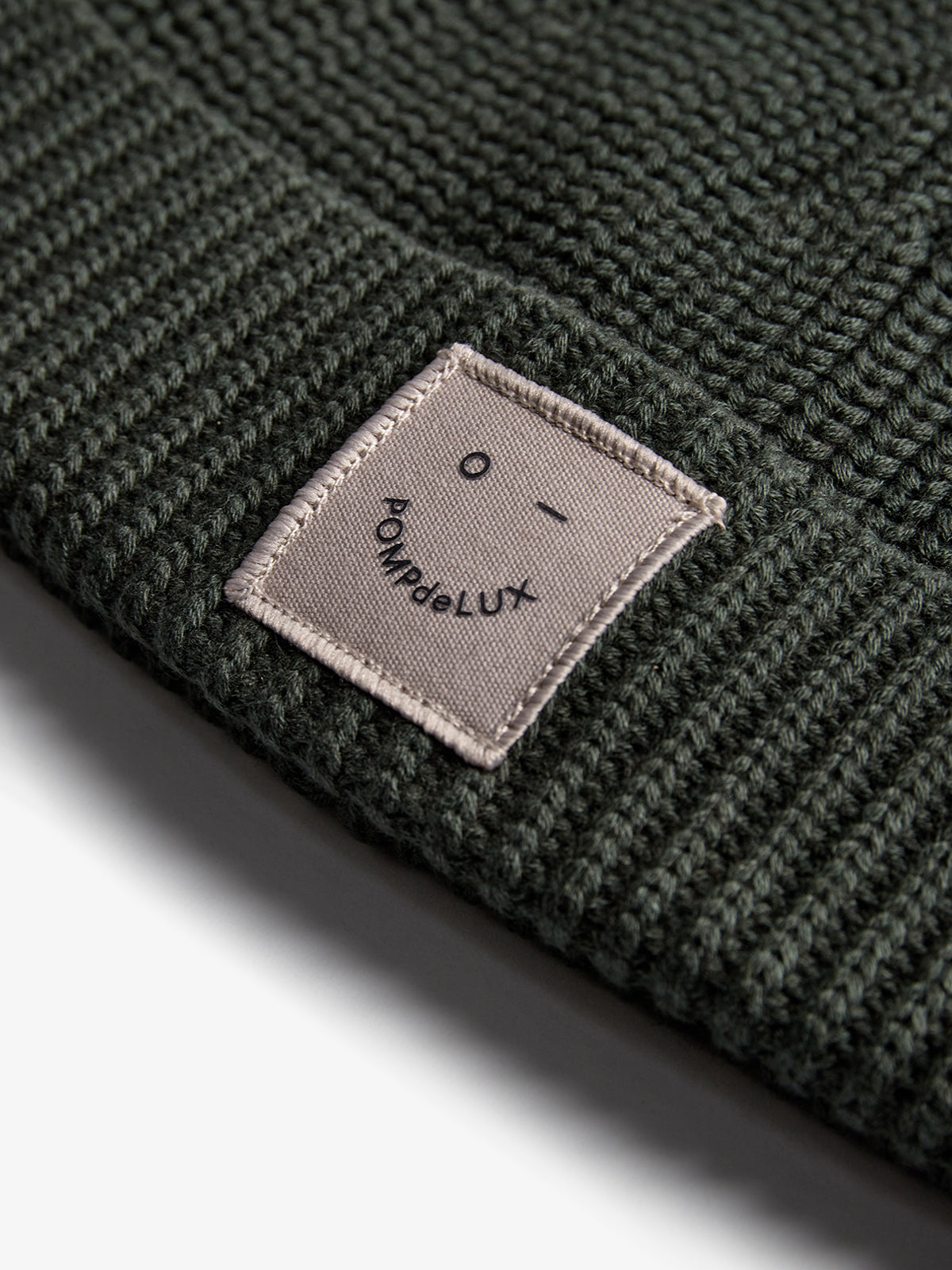 Eine dunkelgrüne Strickmütze zeigt ein aufgenähtes Etikett mit einem lächelnden Gesicht und dem Text "POMPdeLUX". Es liegt auf einer hellen Oberfläche.