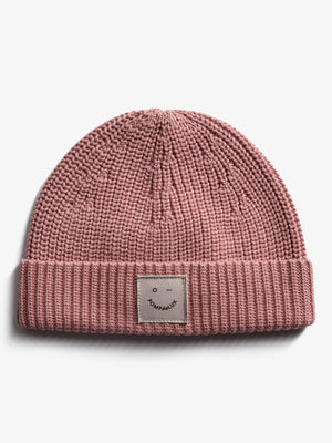 Eine rosa Strickmütze liegt flach auf weißem Hintergrund. Auf der Vorderseite ist ein Etikett mit einem lächelnden Gesicht und der Aufschrift „POMPdeLUX“.