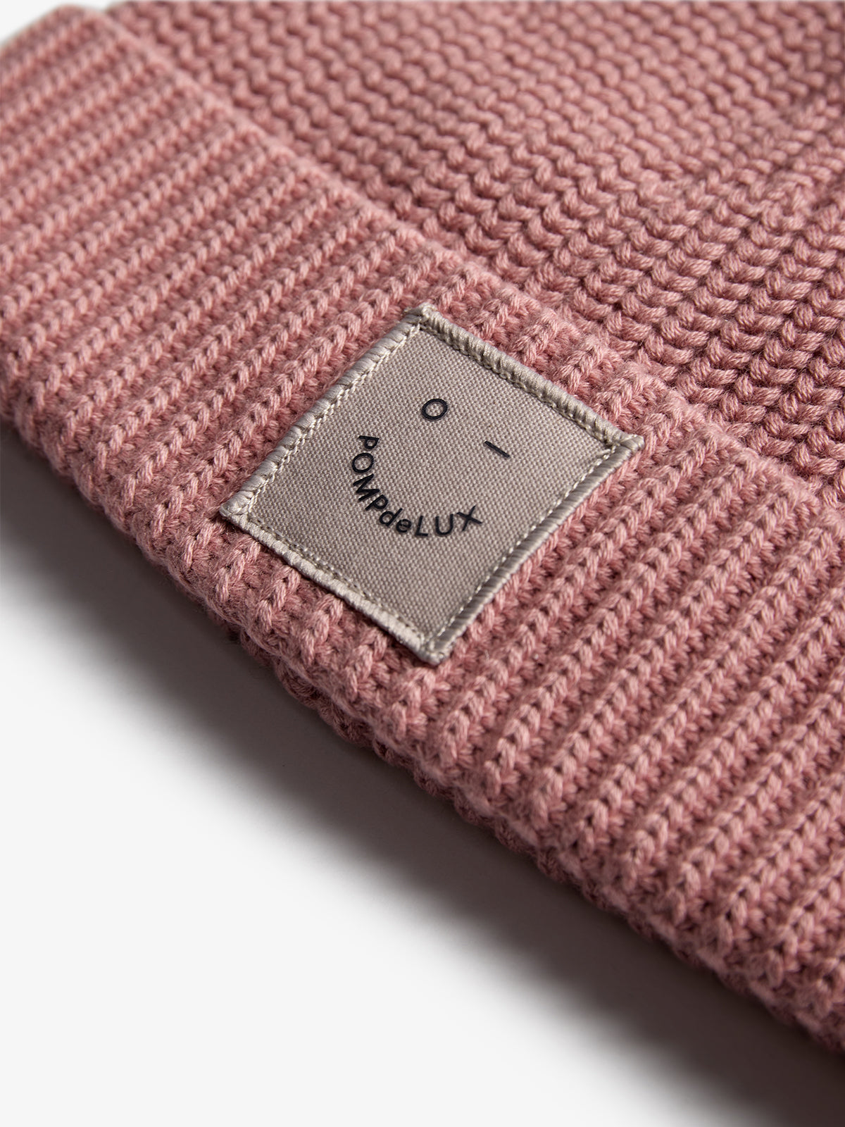Eine rosafarbene Strickmütze liegt auf einer weißen Fläche. Auf der Mütze befindet sich ein Stoffetikett mit dem Text: „O - POMPdeLUX“.