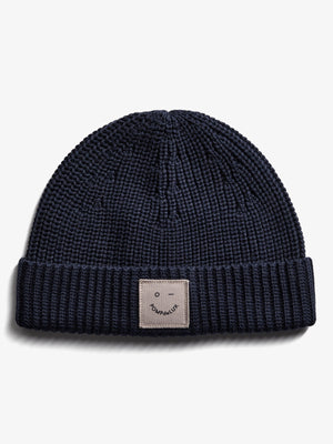 Eine dunkelblaue Strickmütze liegt flach auf weißem Hintergrund. Der umgeschlagene Rand zeigt ein quadratisches Etikett mit einem minimalistischen Gesicht und dem Text "POMPdeLUX".