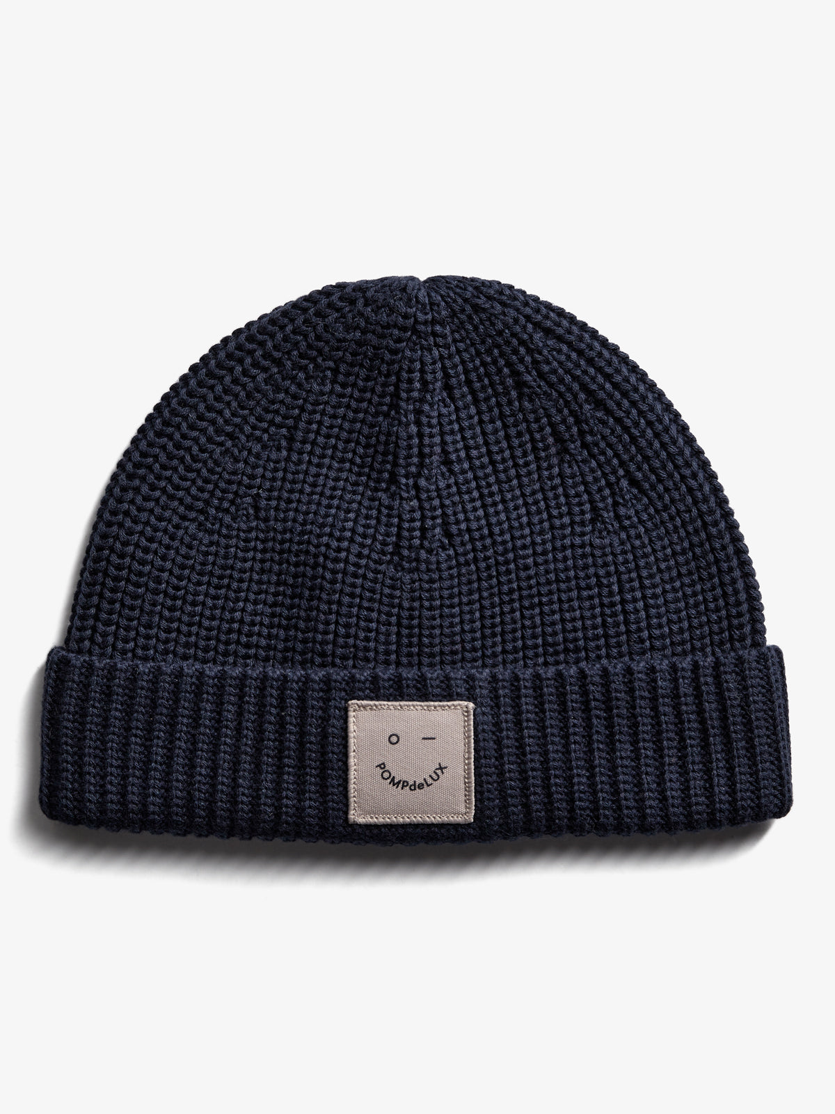 Eine dunkelblaue Strickmütze liegt flach auf weißem Hintergrund. Der umgeschlagene Rand zeigt ein quadratisches Etikett mit einem minimalistischen Gesicht und dem Text "POMPdeLUX".
