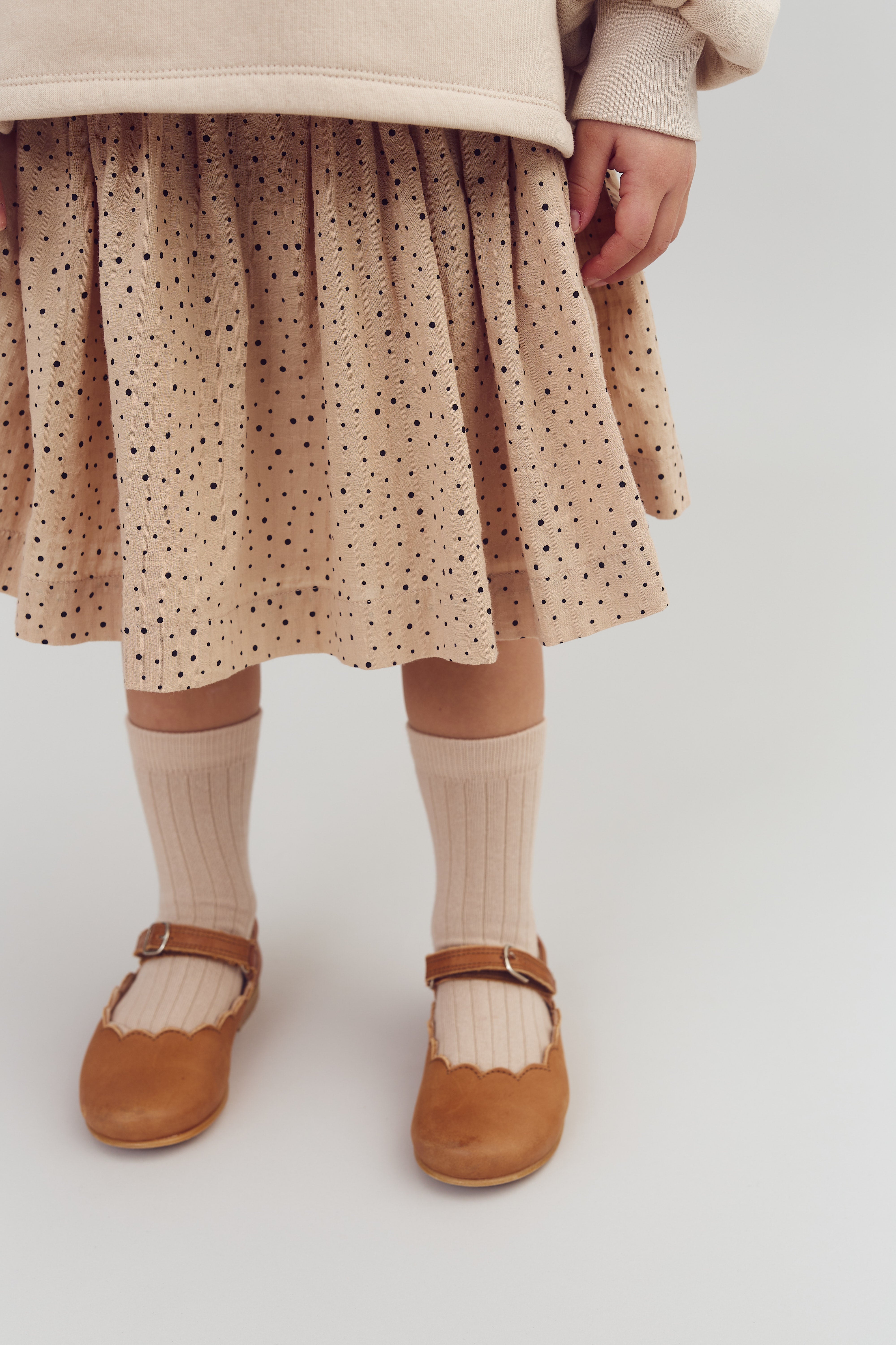 Ein Kind trägt einen gepunkteten Rock und braune Mary-Jane-Schuhe mit weißen Socken. Das Outfit ist in einem minimalistischen, hellen Raum abgebildet.