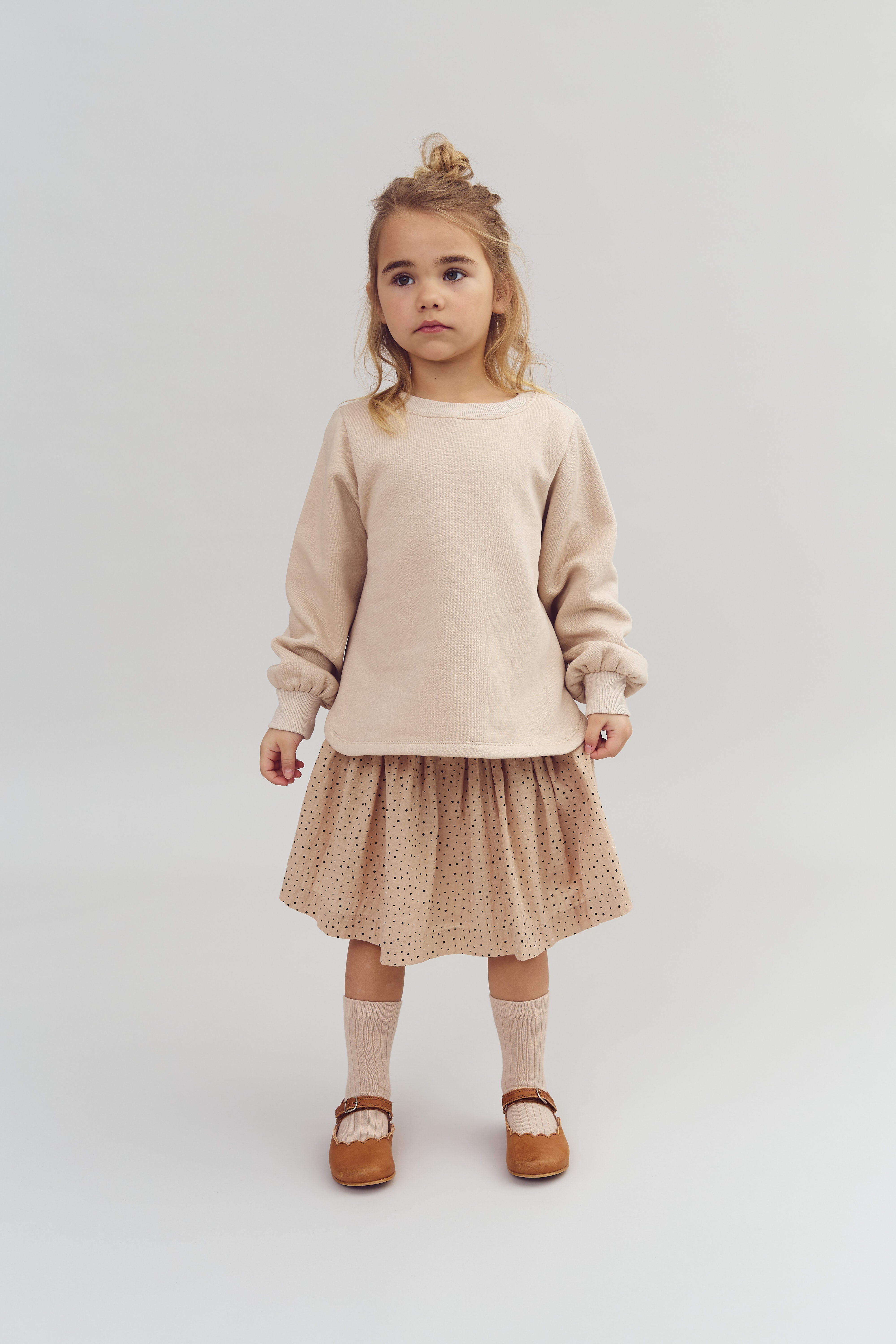 Mädchen steht unbeteiligt im Raum, trägt beigen Pullover, gepunkteten Rock, lange Socken und braune Schuhe. Heller, minimalistischer Hintergrund.