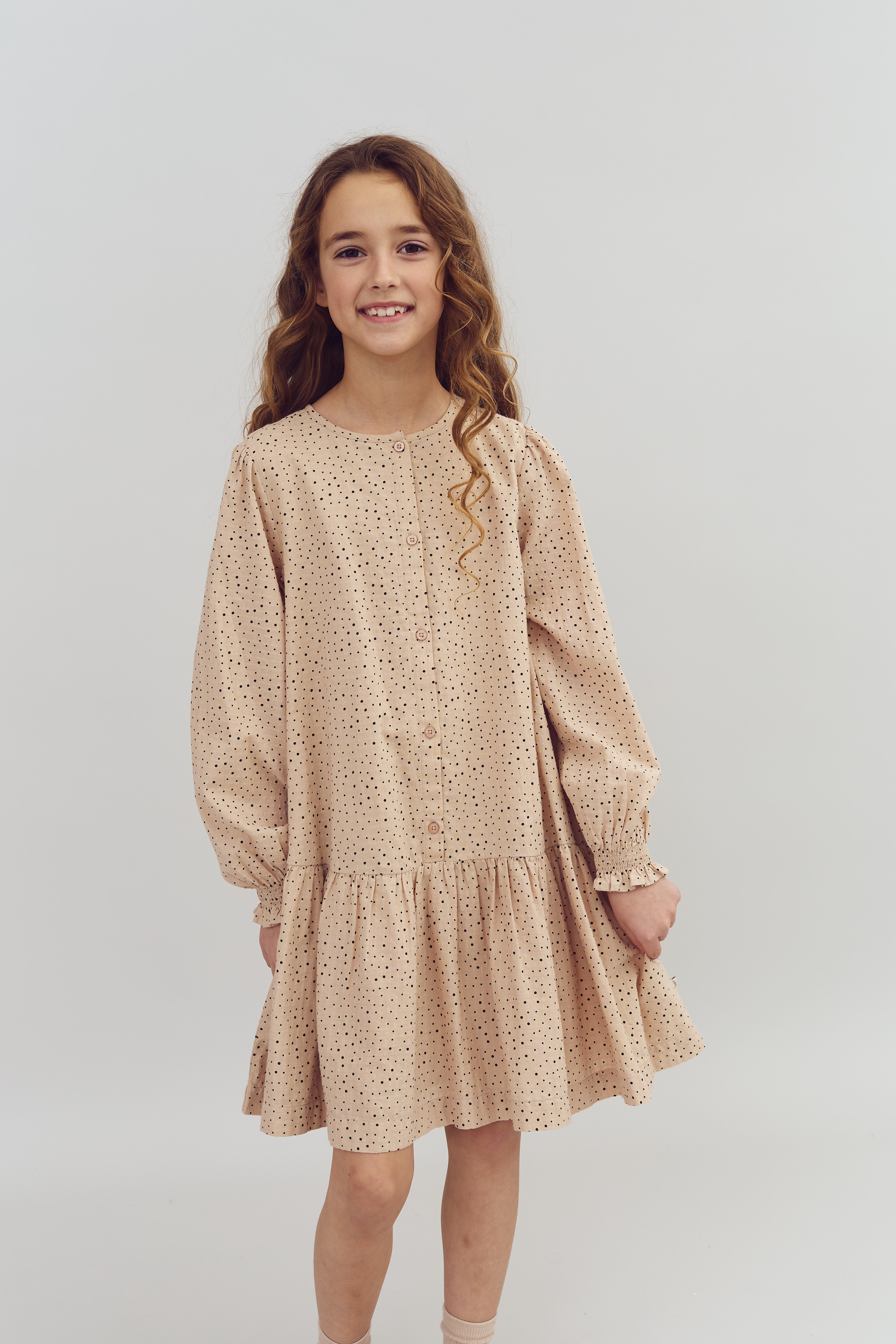 Mädchen trägt ein langärmliges, gepunktetes Kleid und steht lächelnd. Der Hintergrund ist schlicht und hell, was den Fokus auf das Mädchen lenkt.