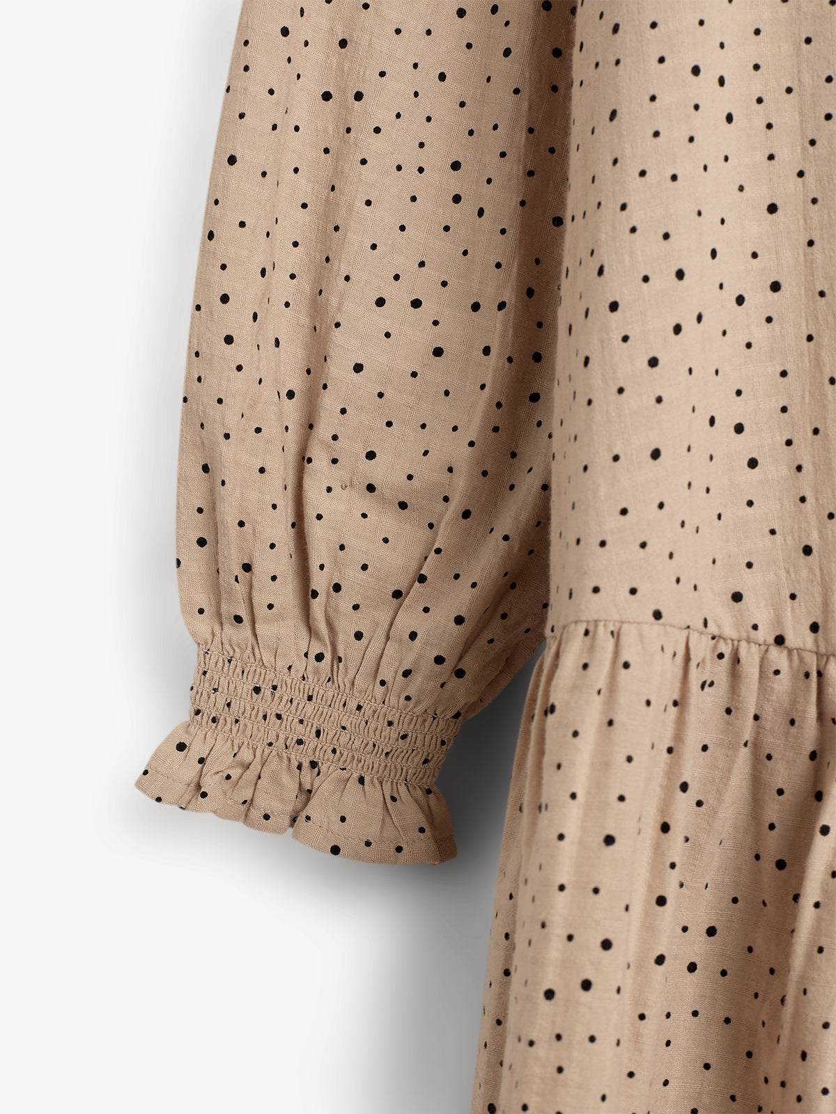 Ein beigefarbenes Kleid mit schwarzen Punkten zeigt eine geraffte, elastische Manschette. Der ärmellose Schnitt und das fließende Material verleihen dem Kleid ein elegantes Aussehen in neutraler Umgebung.