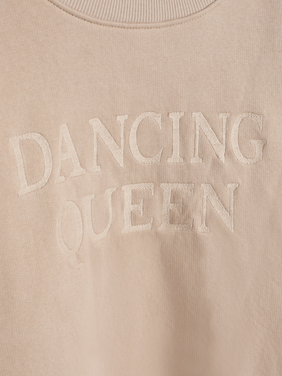 Beiger Pullover mit gesticktem Text "DANCING QUEEN" in großen Buchstaben, aufgenommen aus nächster Nähe.