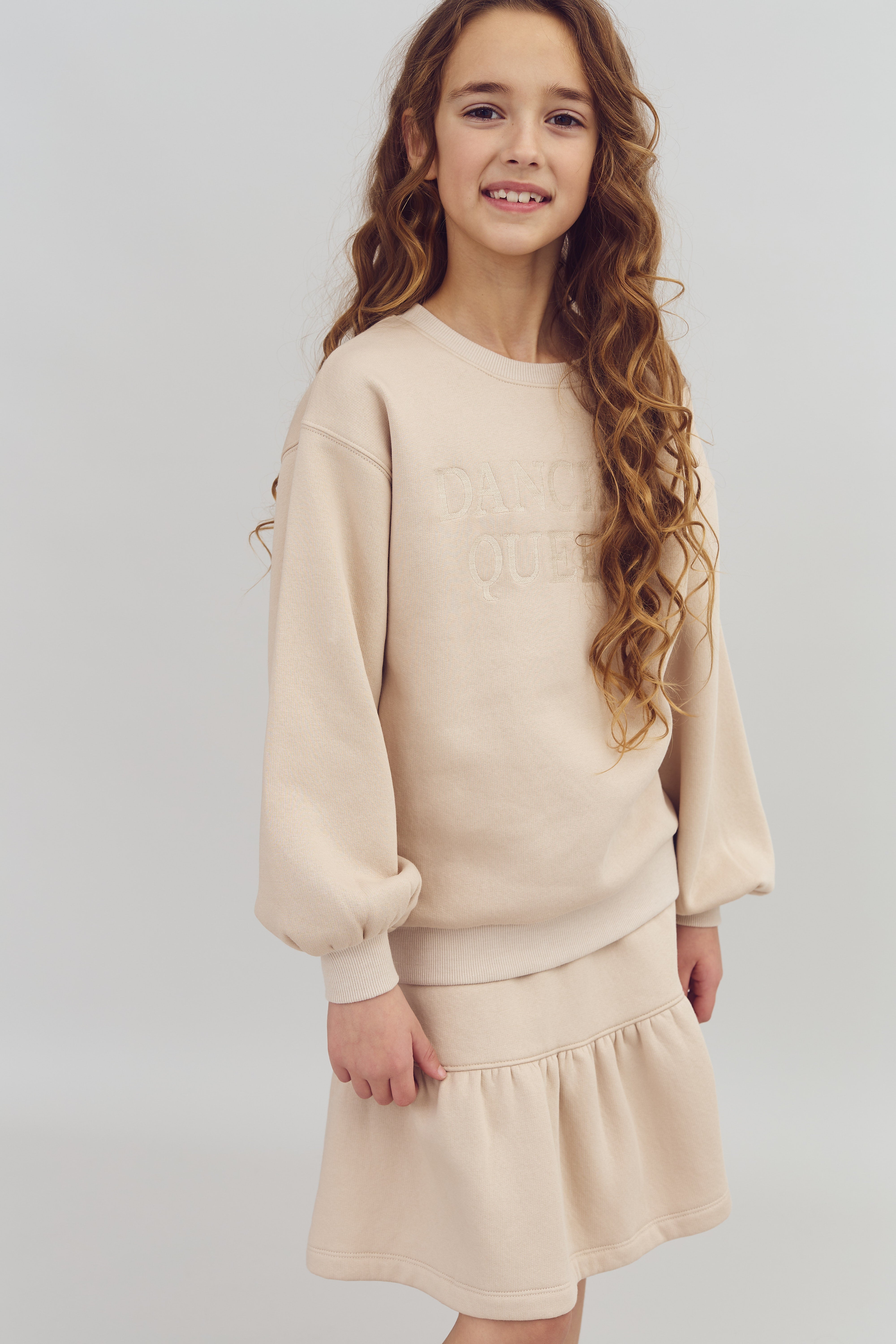 Mädchen steht lächelnd; trägt beigefarbenes Sweatshirt mit Aufschrift "DANCE QUEEN" und passendem ausgestelltem Rock; Hintergrund ist schlicht grau.