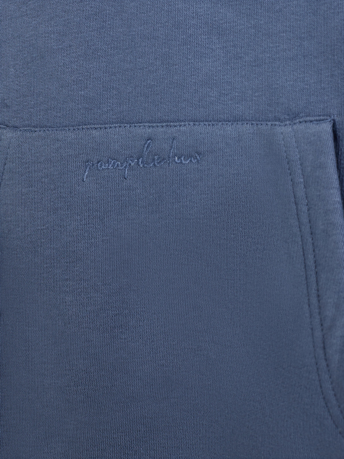 Eine blaue Kapuzenpullover-Tasche mit gesticktem Text "pamplona" in einer strukturierten Stoffoberfläche, aufgenommen in einer Nahaufnahme, zeigt die Nahtdetails und einen schlichten Hintergrund.