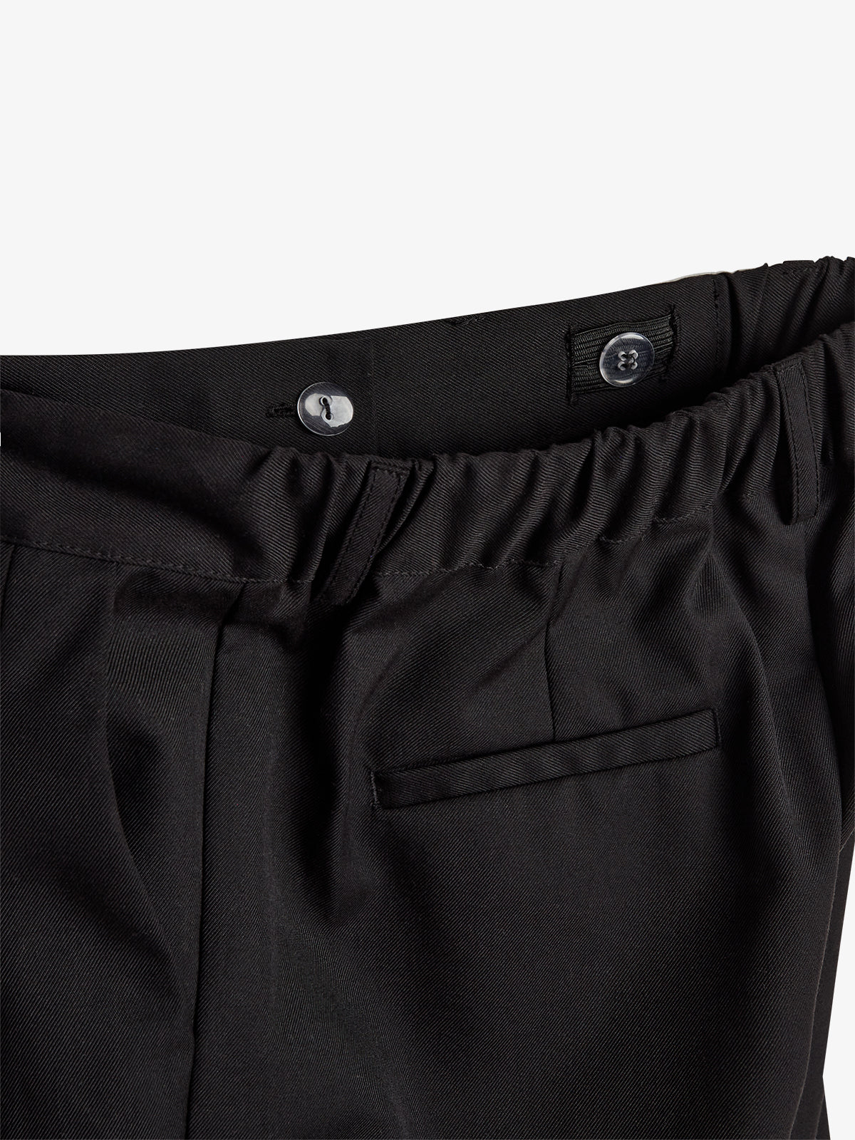 Schwarze Hose, liegend, mit elastischem Bund und einem Knopfverschluss. Im Hintergrund ist ein neutraler, heller Hintergrund zu sehen. Eine Tasche ist erkennbar.