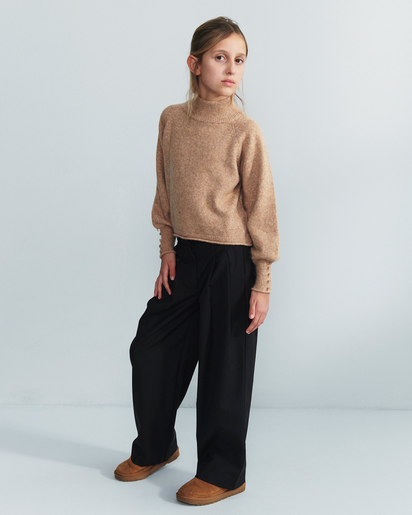 Ein Kind steht ruhig, trägt einen braunen Rollkragenpullover, schwarze weite Hosen und braune Schuhe. Der Hintergrund ist einfarbig hellgrau.