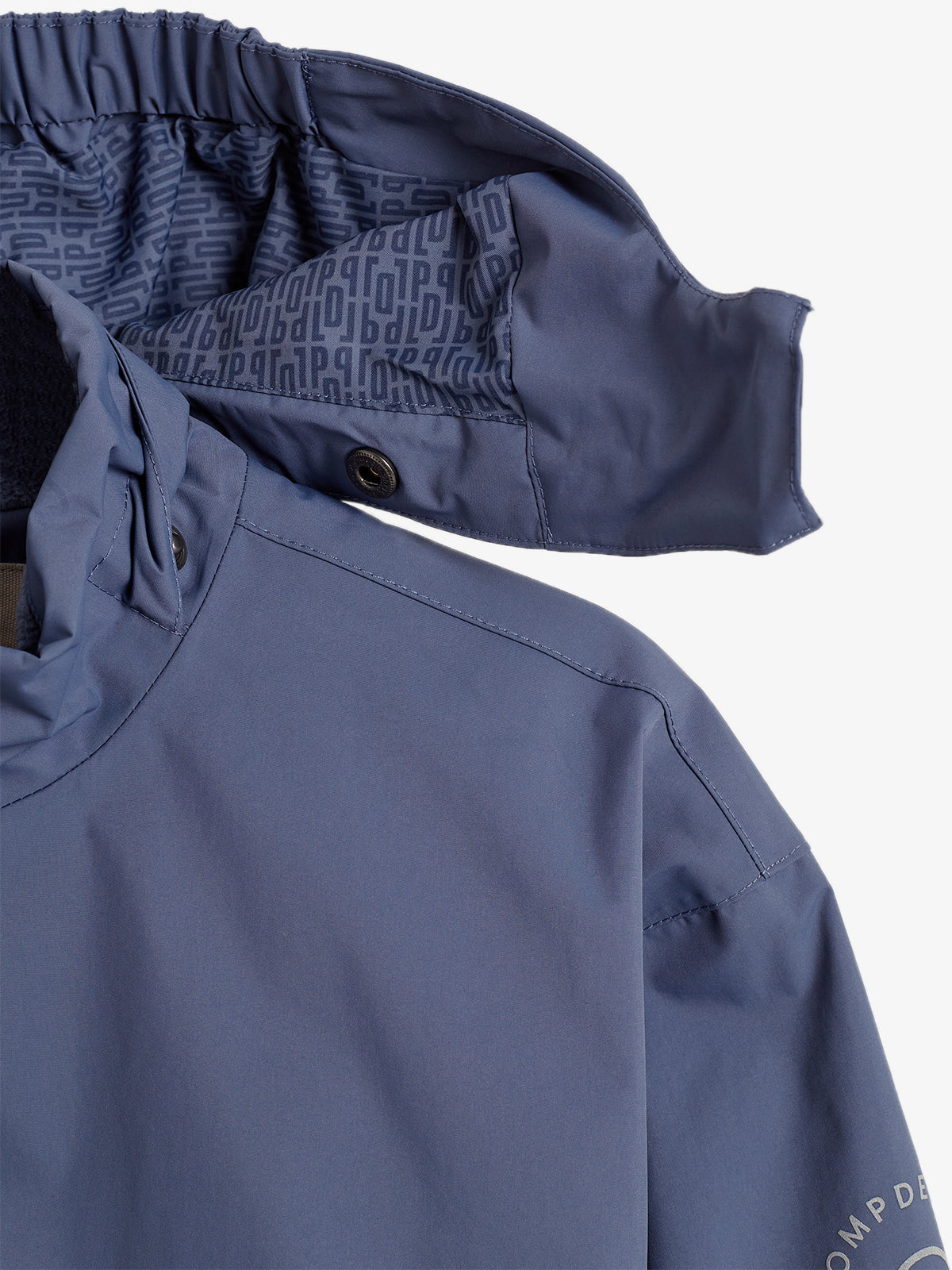Eine blaue Jacke mit einer Kapuze zeigt ein dezentes Muster im Inneren. Knöpfe befestigen den Kapuzenrand, während am Ärmel die teilweise sichtbare Aufschrift "COMP DE" erscheint.