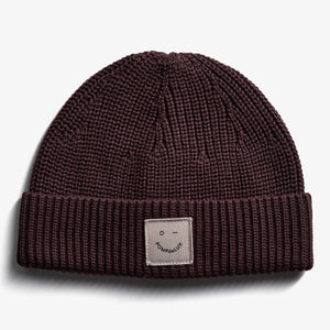 Eine dunkelbraune Strickmütze liegt auf einem weißen Hintergrund. Auf der Vorderseite ist ein beige Etikett mit einem Gesicht und dem Text "POMPdeLUX" aufgenäht.