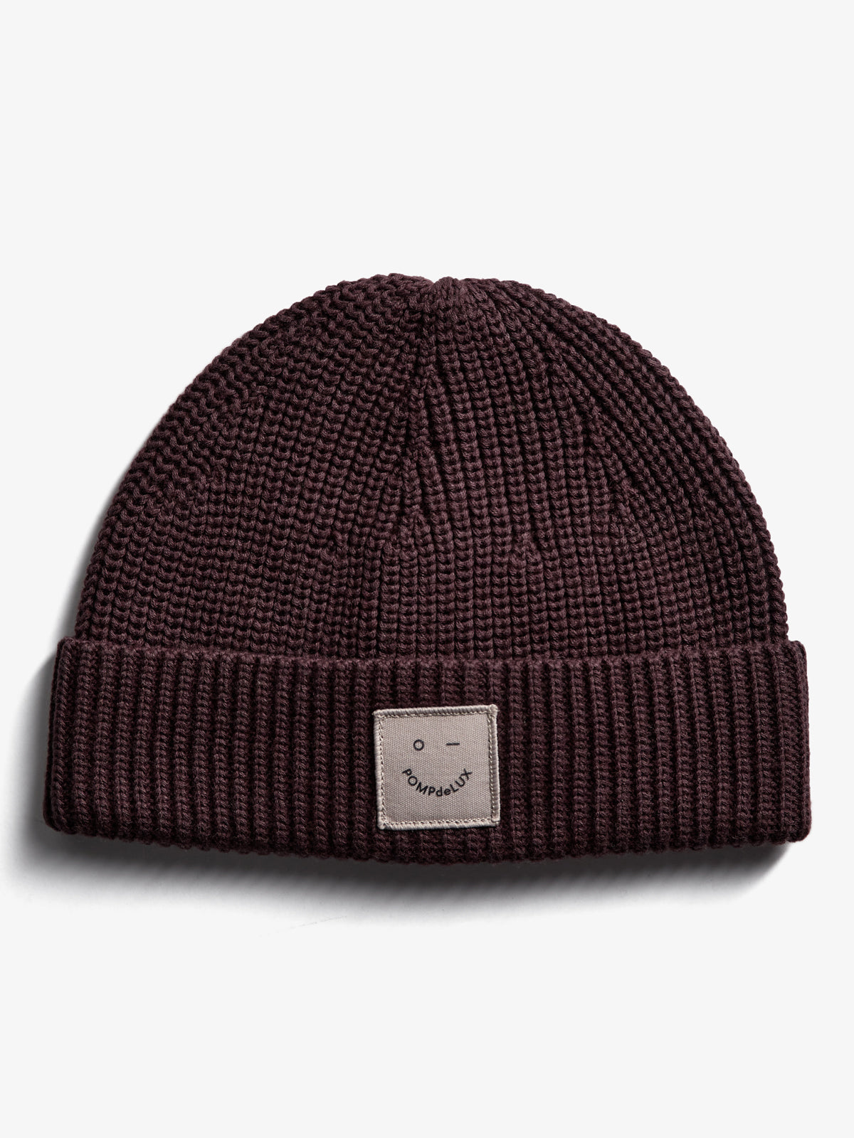 Eine dunkelbraune Strickmütze liegt auf einem weißen Hintergrund. Auf der Vorderseite ist ein beige Etikett mit einem Gesicht und dem Text "POMPdeLUX" aufgenäht.