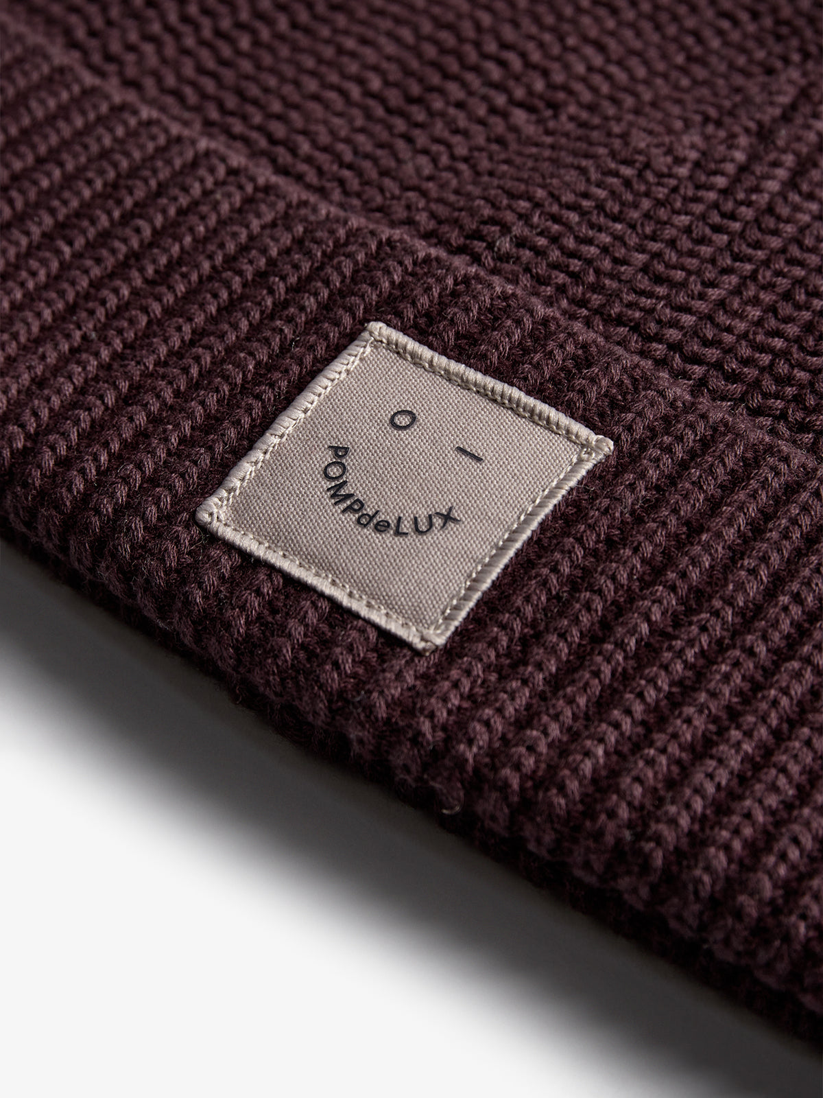 Braune Strickmütze mit rechteckigem Etikett, das ein minimalistisches Gesicht und den Text "POMPdeLUX" zeigt. Die Mütze liegt auf einer glatten, hellen Oberfläche.
