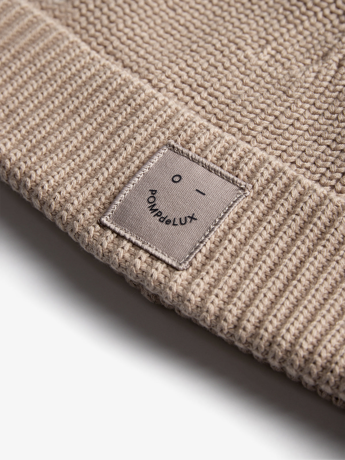 Eine cremefarbene Strickmütze mit einem rechteckigen Etikett trägt ein minimalistisches Gesicht und den Text "POMPdeLUX". Liegt auf einer weißen Oberfläche.